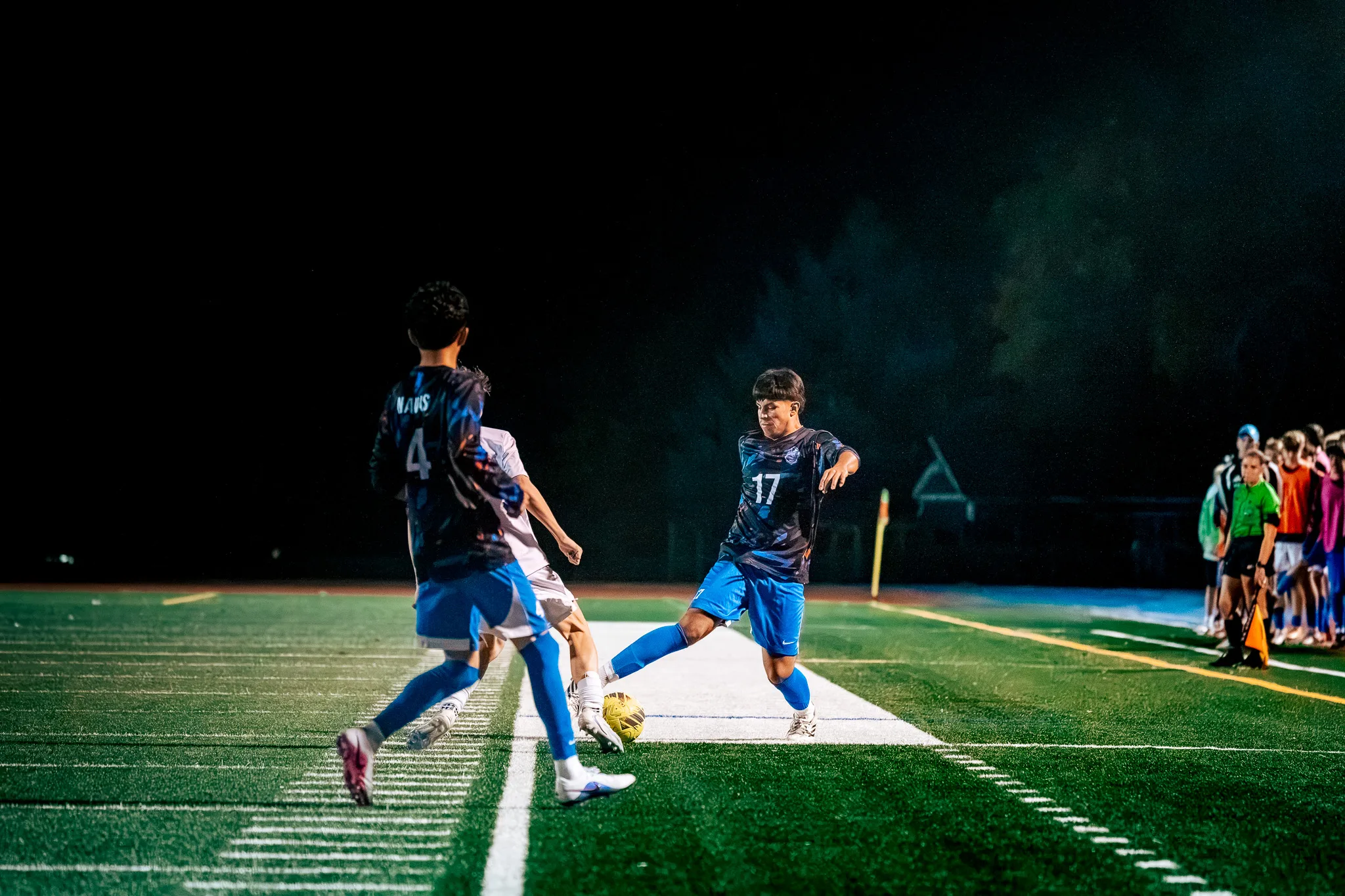 Centaurus number 17 hits the ball along the touchline as his teammate number 4 and a Mead player scramble in front of him, with an assistant referee in green and a row of players at the right edge