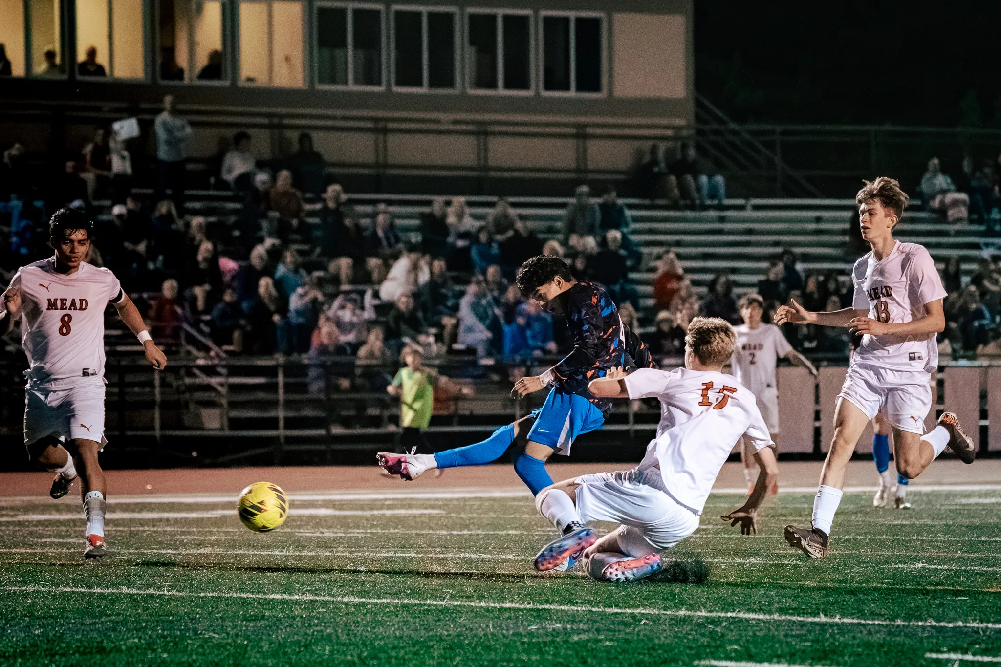 A Centaurus player strikes the ball past a sliding Mead number 15 as Mead numbers 8 and 13 run in and spectators watch from the bleachers