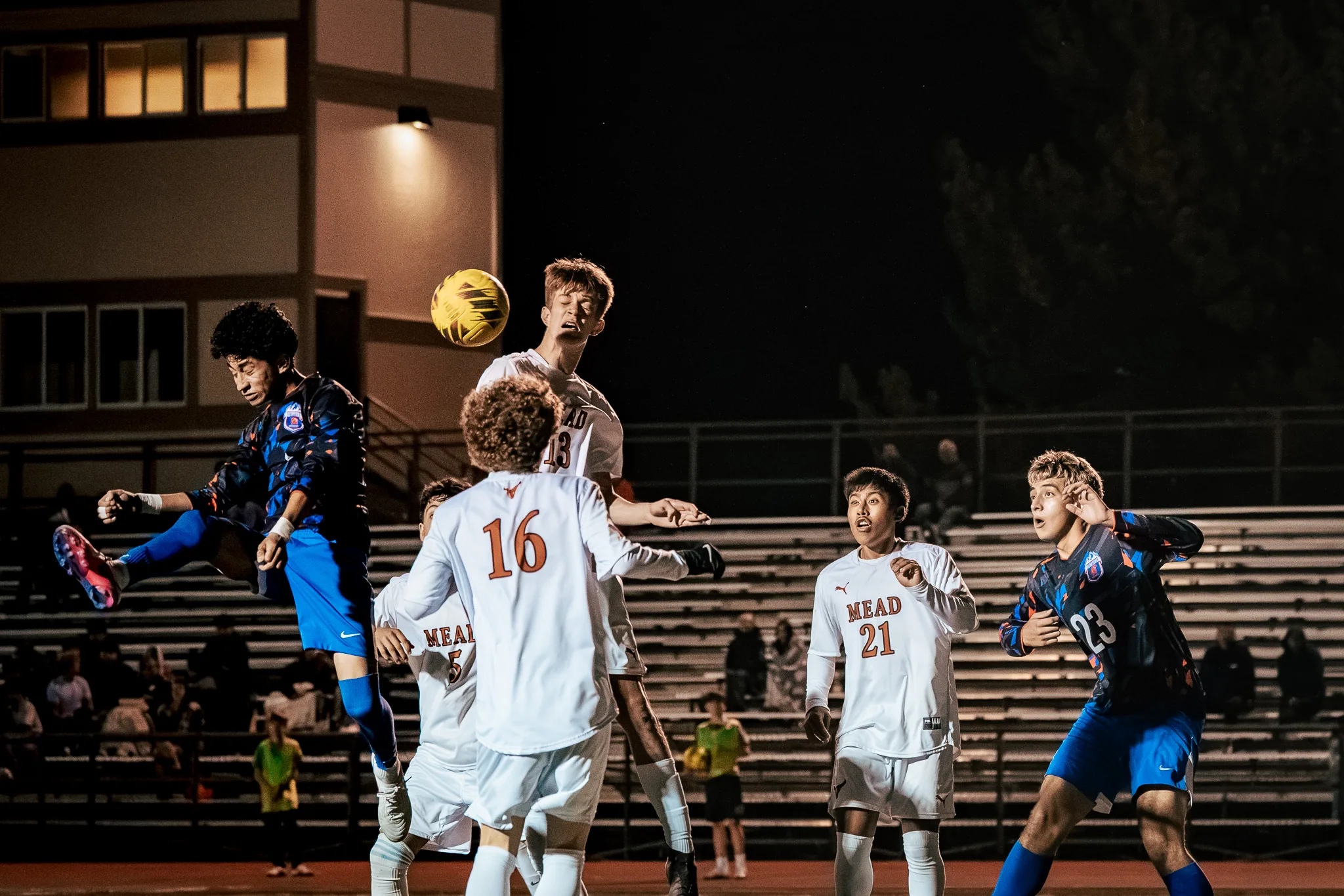 A Centaurus player and Mead number 13 leap for a high ball in front of the stands as Mead number 16 crowds in from the foreground and Mead number 21 and Centaurus number 23 watch from the right