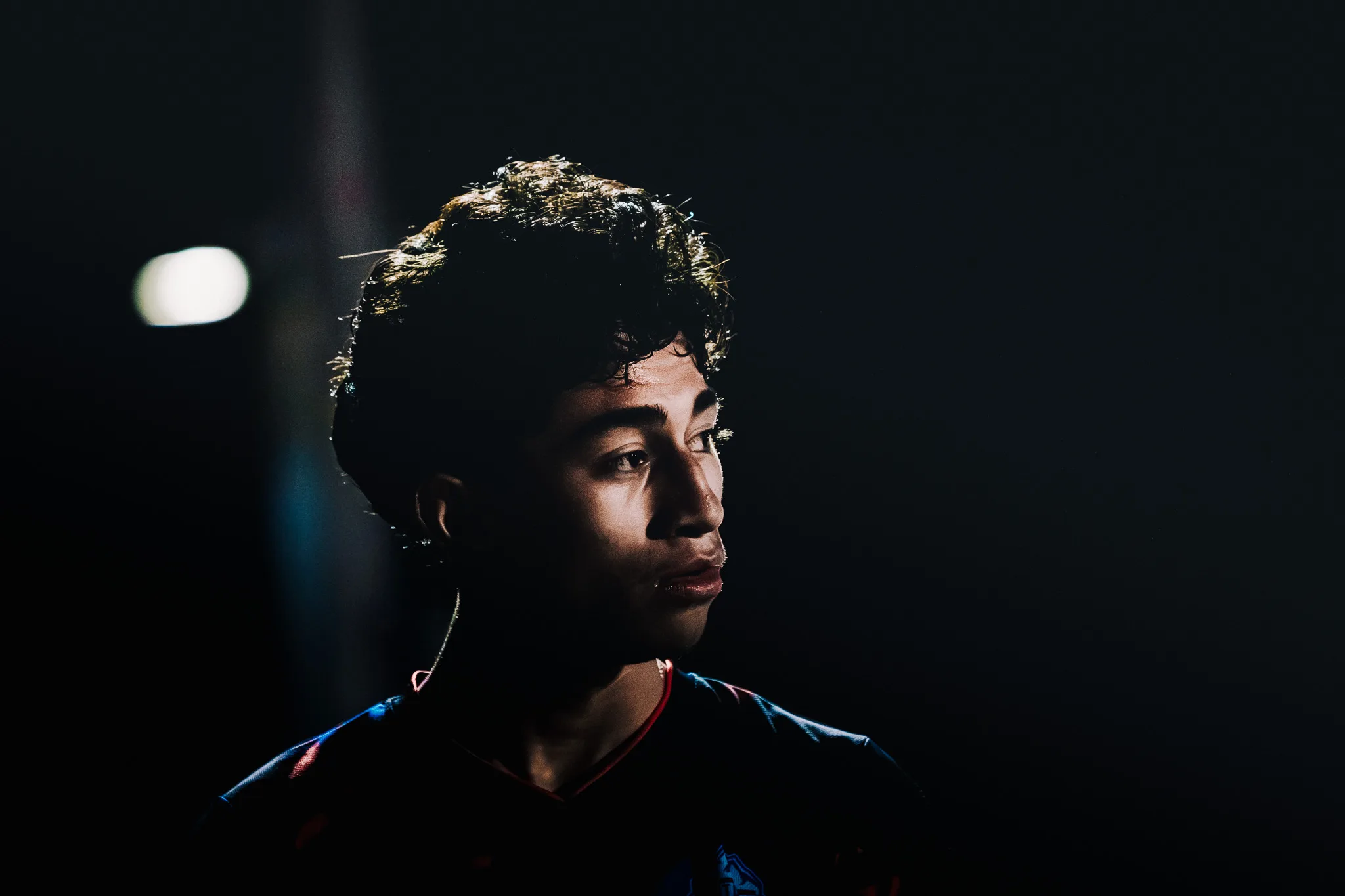 A Centaurus player in profile, his face lit by a thin band of light against the black night, a stadium light glowing out of focus at the left
