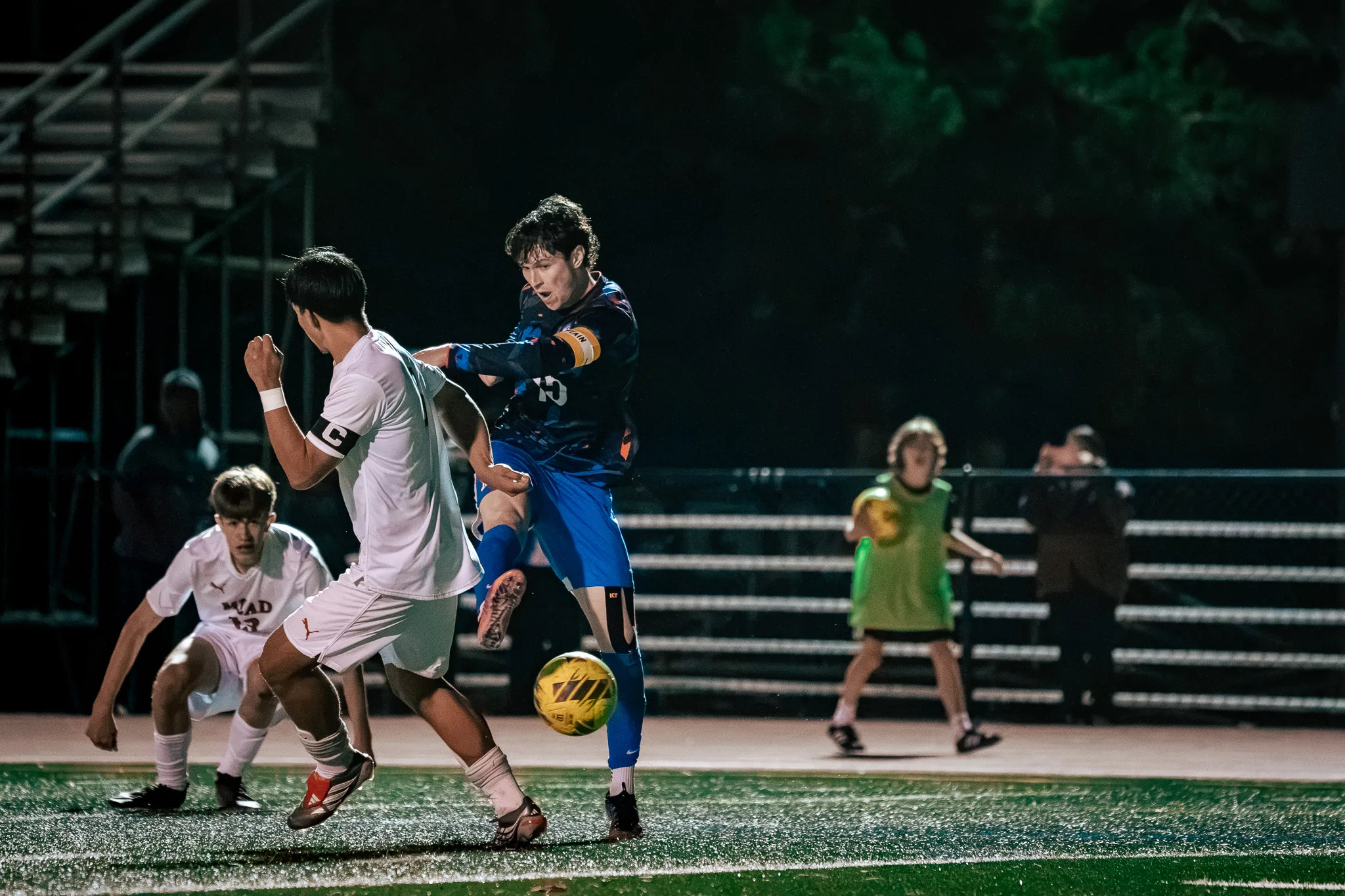 Centaurus number 15, in a captain's armband, strikes the ball past a Mead player who wears his own captain's armband while another Mead player crouches at the left and a person in a green bib holds a spare ball at the right