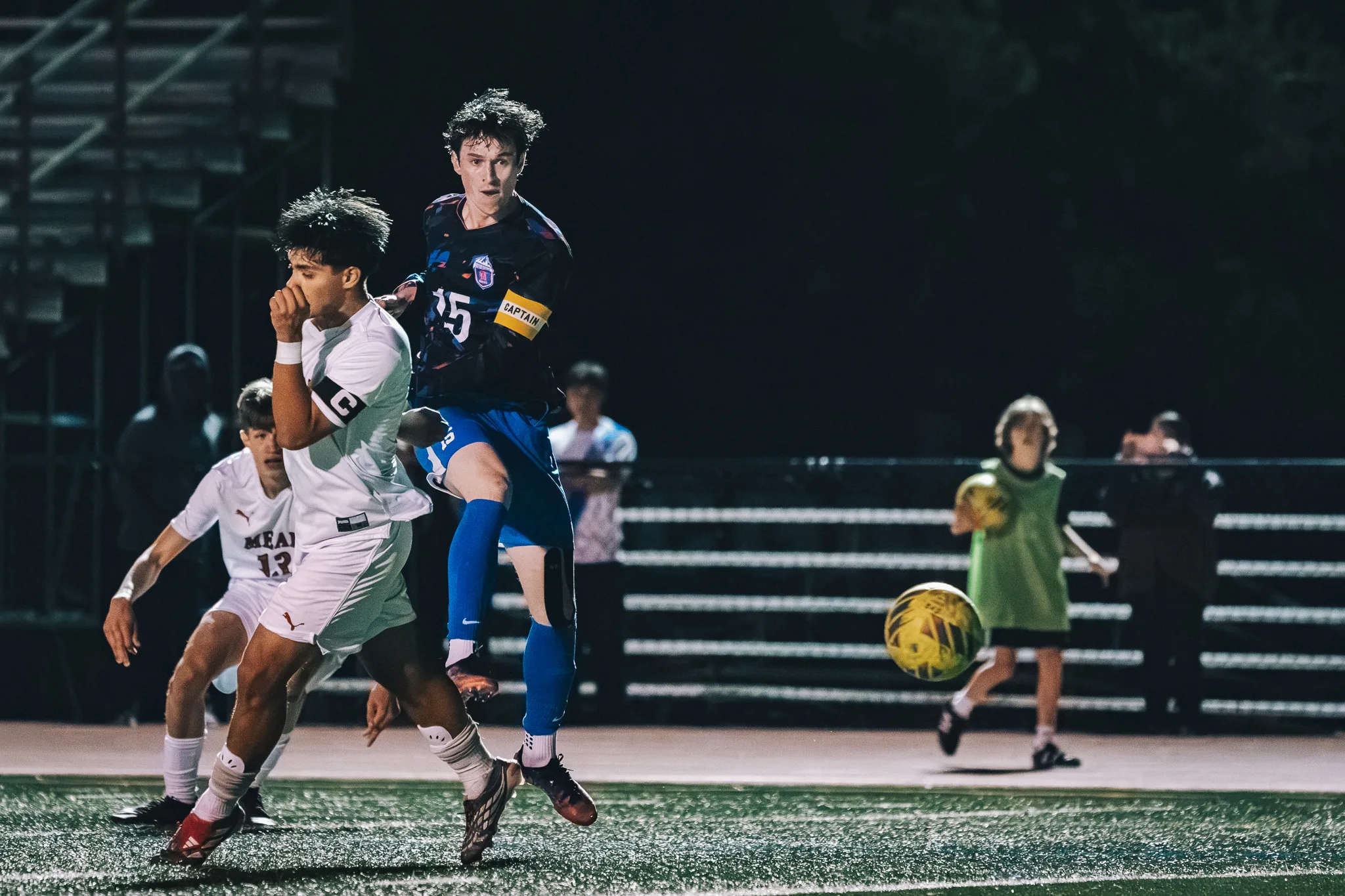 Centaurus number 15, in a captain's armband, follows through on a kick as a Mead player with his own captain's armband covers his face and Mead number 13 runs behind them, a person in a green bib holding a spare ball at the right