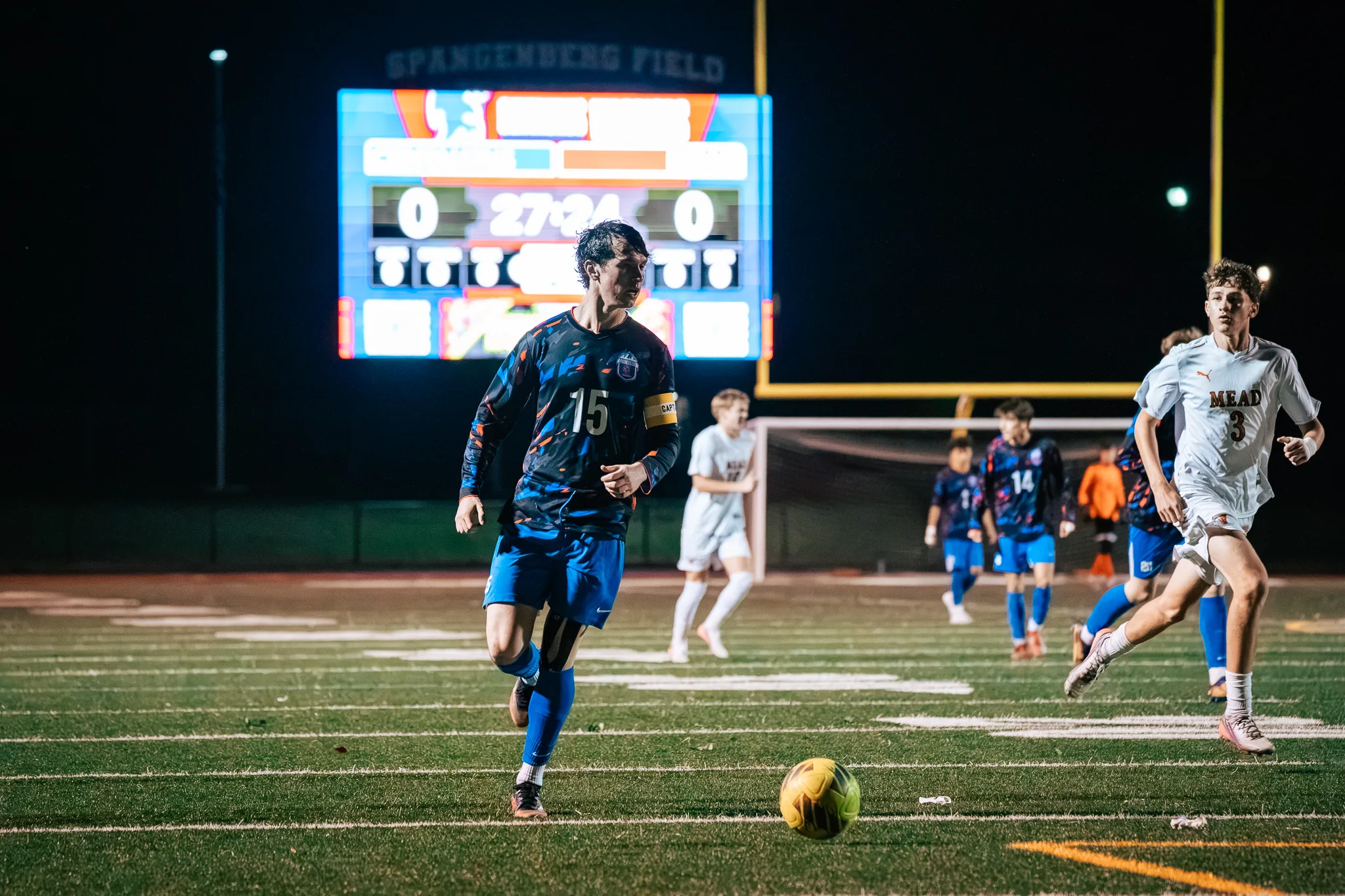 Centaurus number 15, in a captain's armband, runs onto the ball as Mead number 3 sprints in from the right, the scoreboard behind them showing 0 to 0