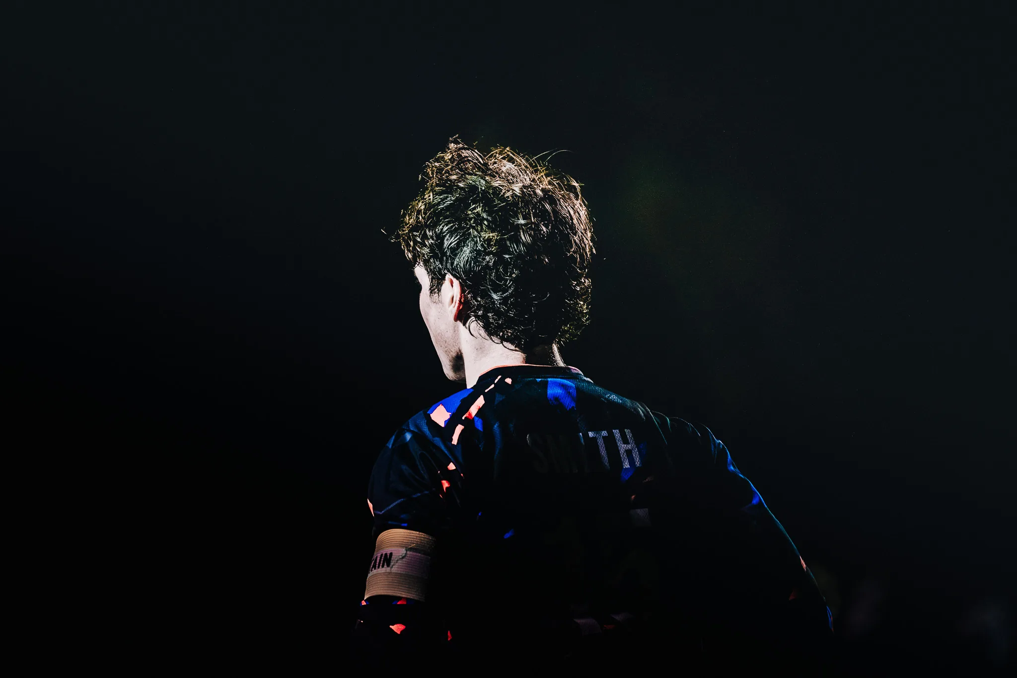 Seen from behind against a black night, a Centaurus player with curly hair and a captain's armband stands lit by a narrow beam of light