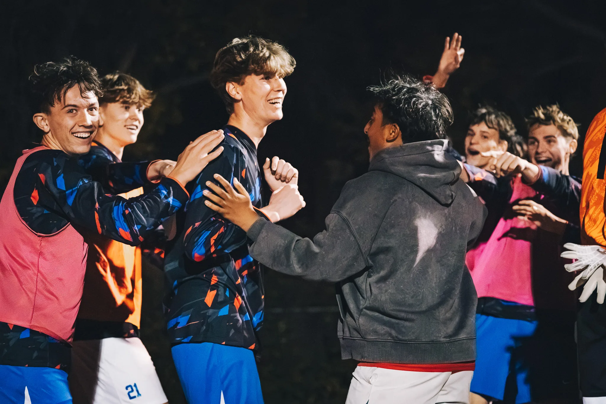 At the sideline, laughing Centaurus players in bibs and jerseys crowd around a teammate, a young man in a gray hoodie reaching toward him, while the goalkeeper's orange jersey and white gloves show at the right edge