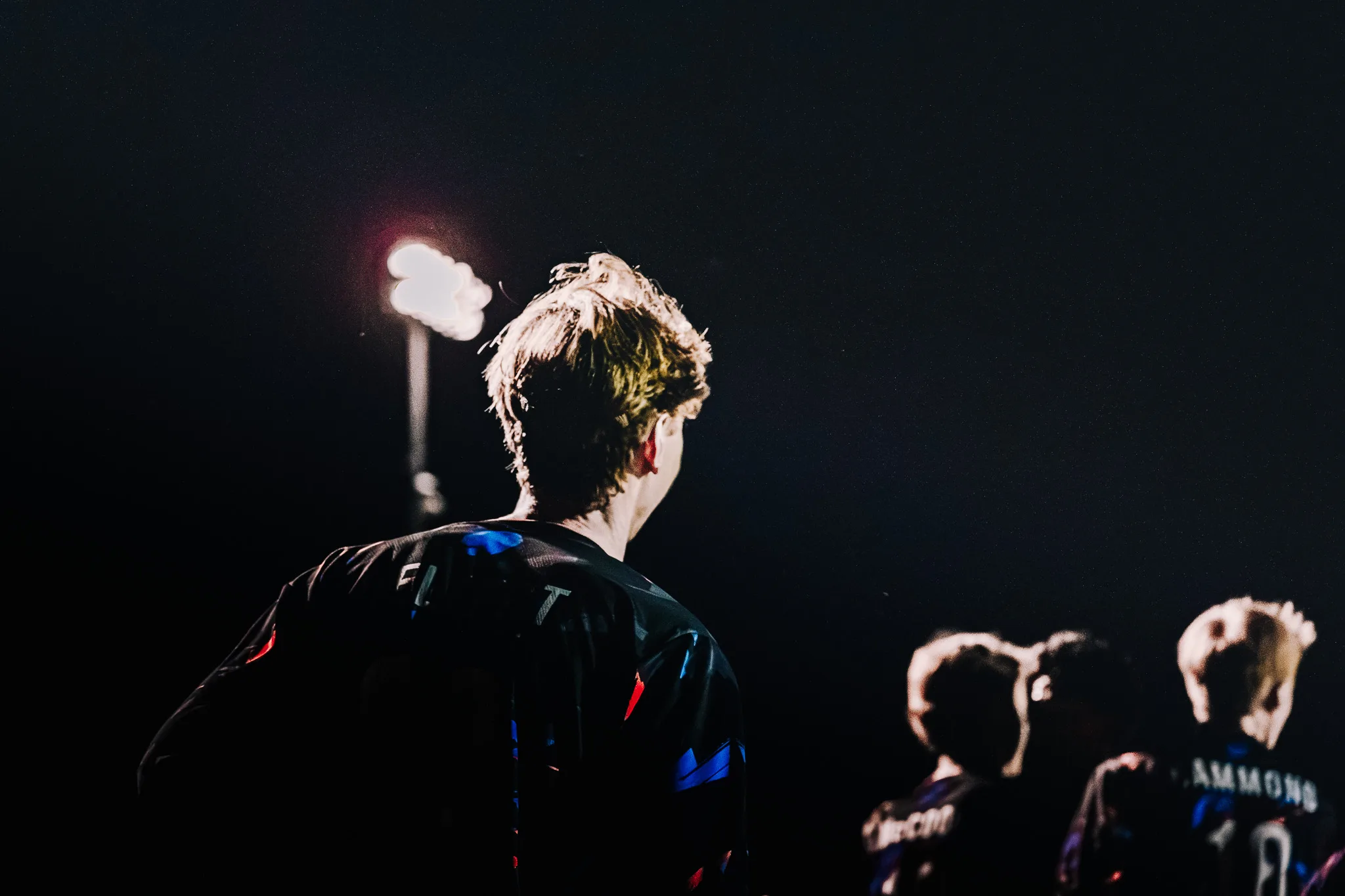 Seen from behind and lit by a thin light, a Centaurus player with tousled hair looks off to the side while three teammates stand blurred at the lower right and a stadium light glows at the left