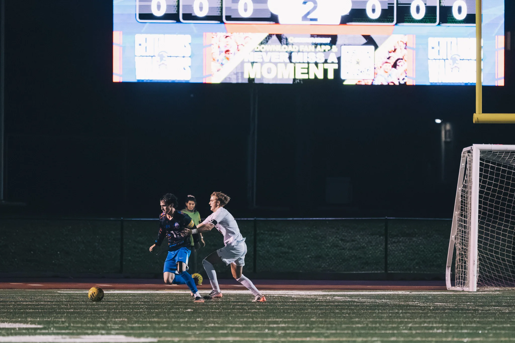 A Mead player grabs at the arm of a Centaurus player as they run along the far side of the field, a ball rolling at the left and the scoreboard glowing above them