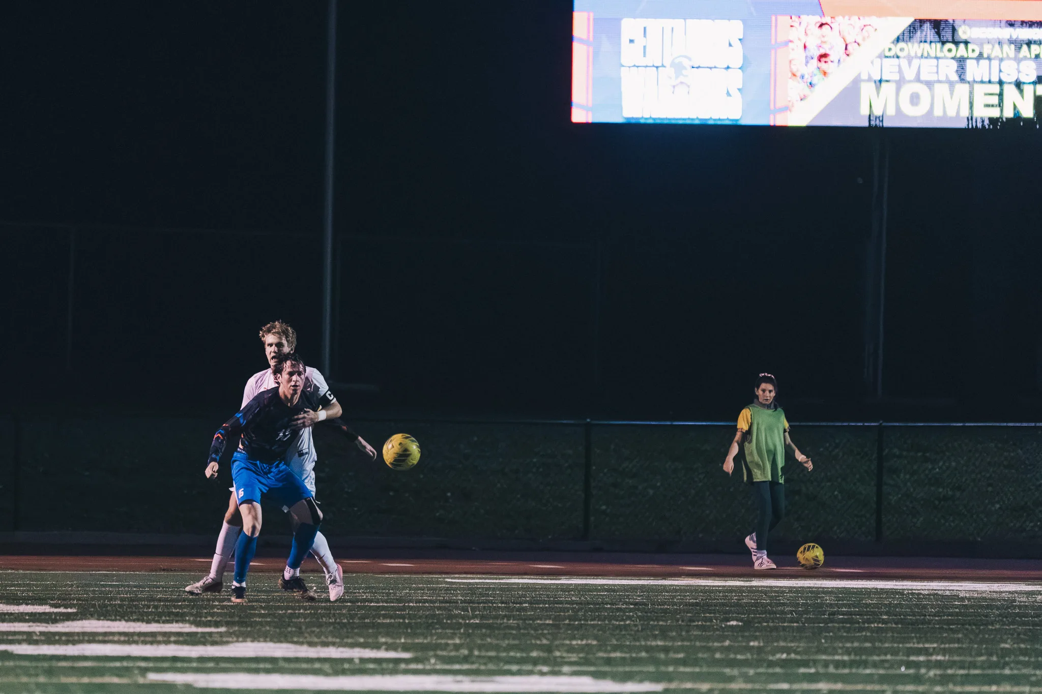 A Mead player stands right behind a Centaurus player with a hand on his back as both watch the ball roll ahead, a person in a green bib at the right