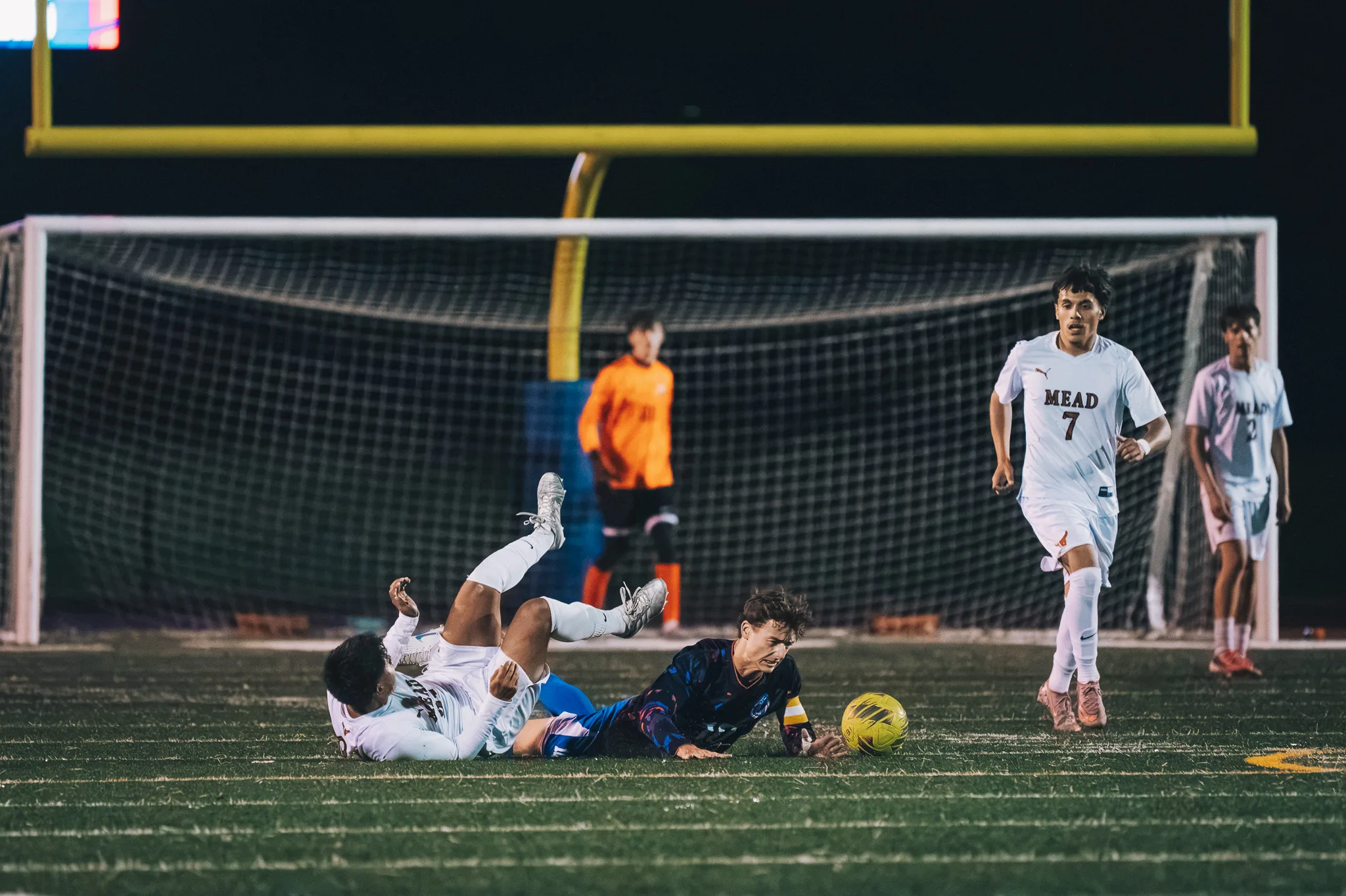 A Mead player lies on his back with a leg raised beside a Centaurus player sprawled on the turf next to the ball, as Mead number 7 runs up and the Centaurus goalkeeper in orange watches from the goal