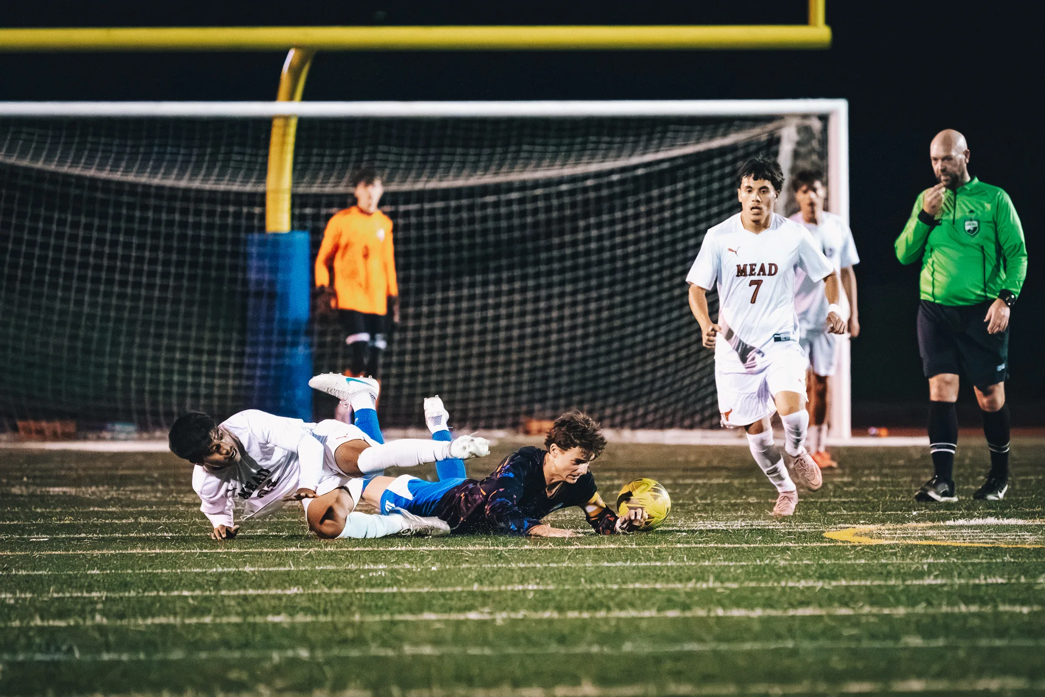A Centaurus player and a Mead player slide to the turf beside the ball as Mead number 7 runs up, a referee in green raises a whistle to his mouth and the Centaurus goalkeeper in orange waits in the goal behind