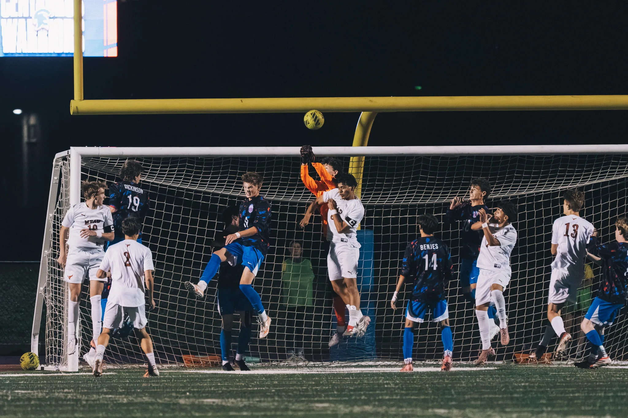 In front of the Centaurus goal the goalkeeper in orange stretches up with both hands as a Mead player in a captain's armband rises beside him, Centaurus numbers 19 and 14 and Mead numbers 2 and 13 around them and the ball above the crossbar