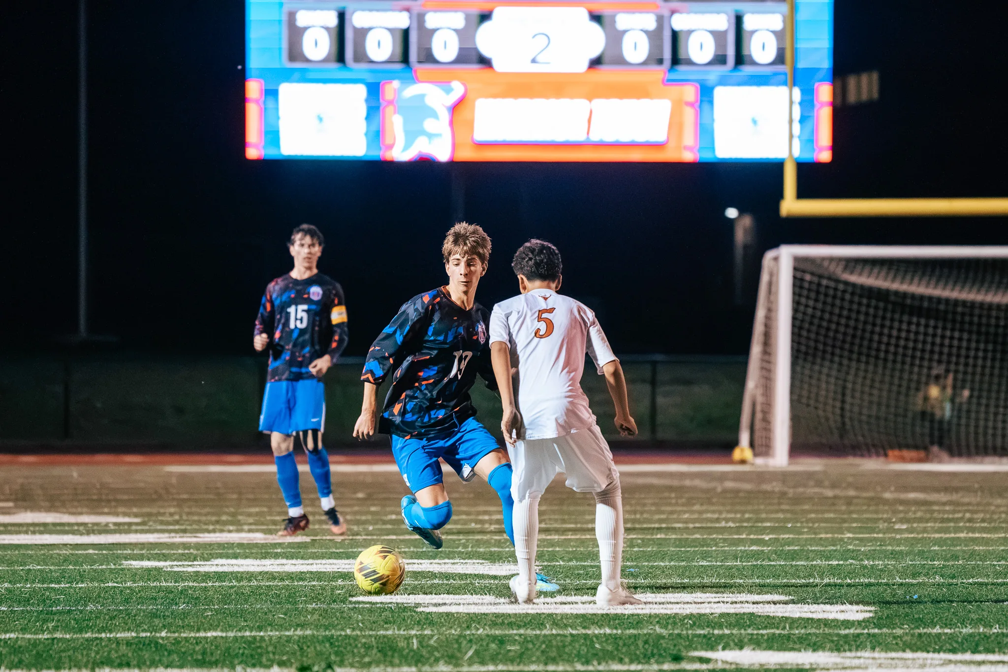 Centaurus number 19 dribbles at Mead number 5 while Centaurus number 15, in a captain's armband, follows behind, the scoreboard and a goal in the background