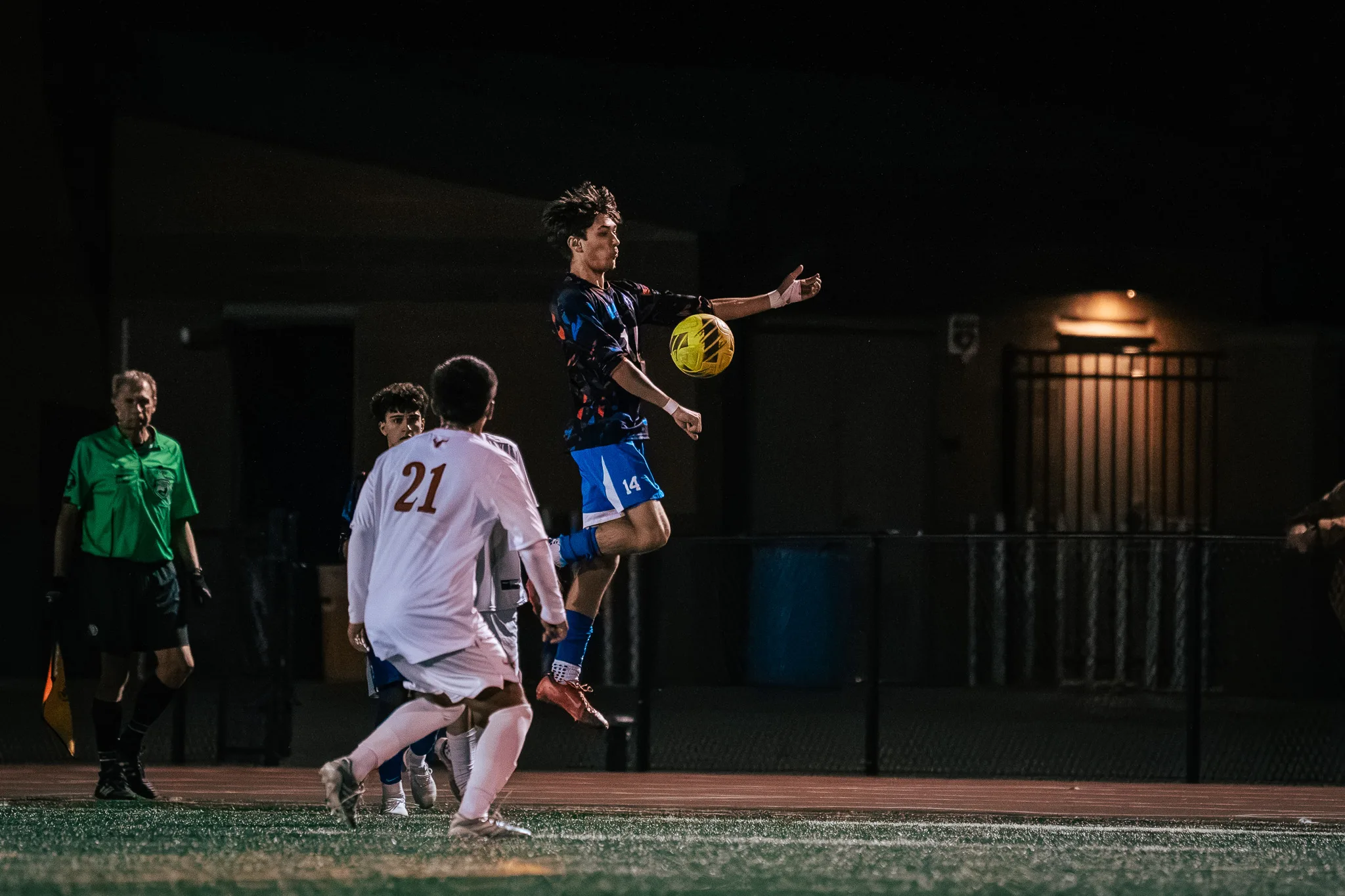Centaurus number 14 leaps with an arm out to control the ball in front of the field house wall as Mead number 21 closes in from the left foreground and an assistant referee in green looks on