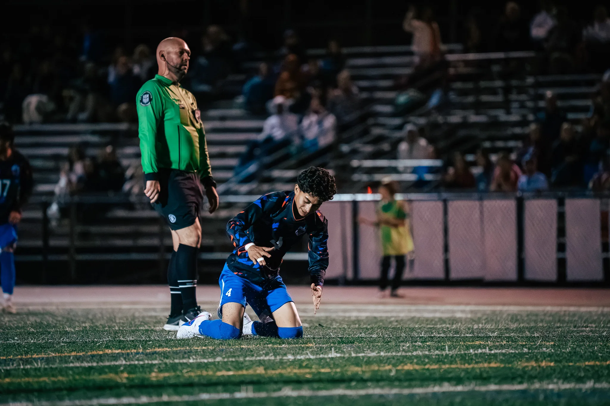 Kneeling on the turf, Centaurus number 4 points at the ground as a referee in a green shirt looks on, spectators in the bleachers behind