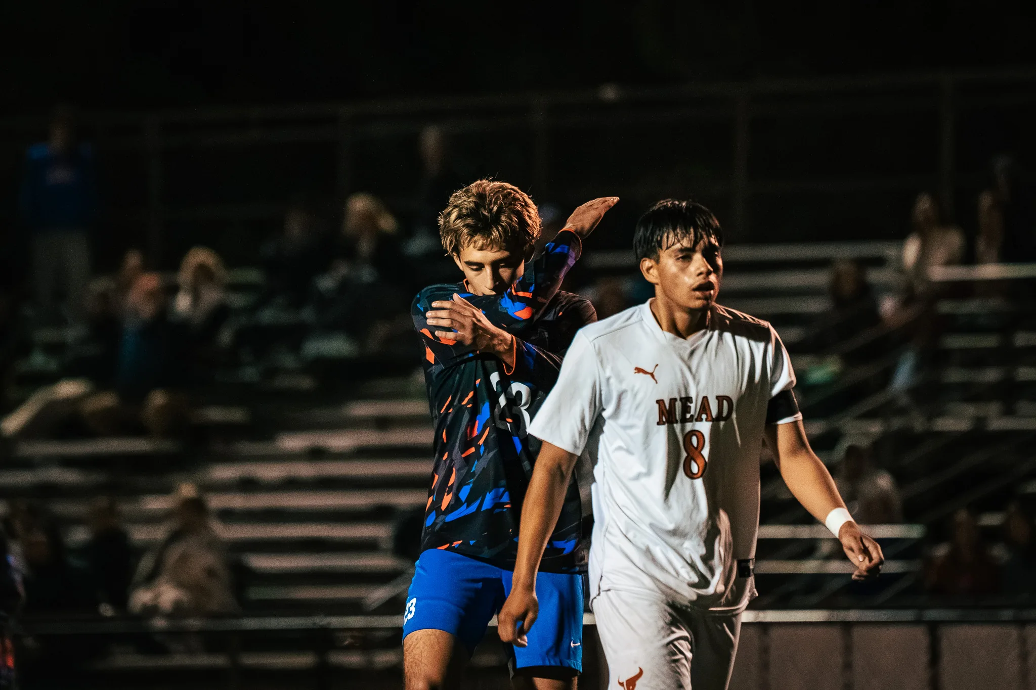 A Centaurus player turns his face into the crook of one raised arm beside Mead number 8, who walks past in the foreground, with spectators in the bleachers behind them