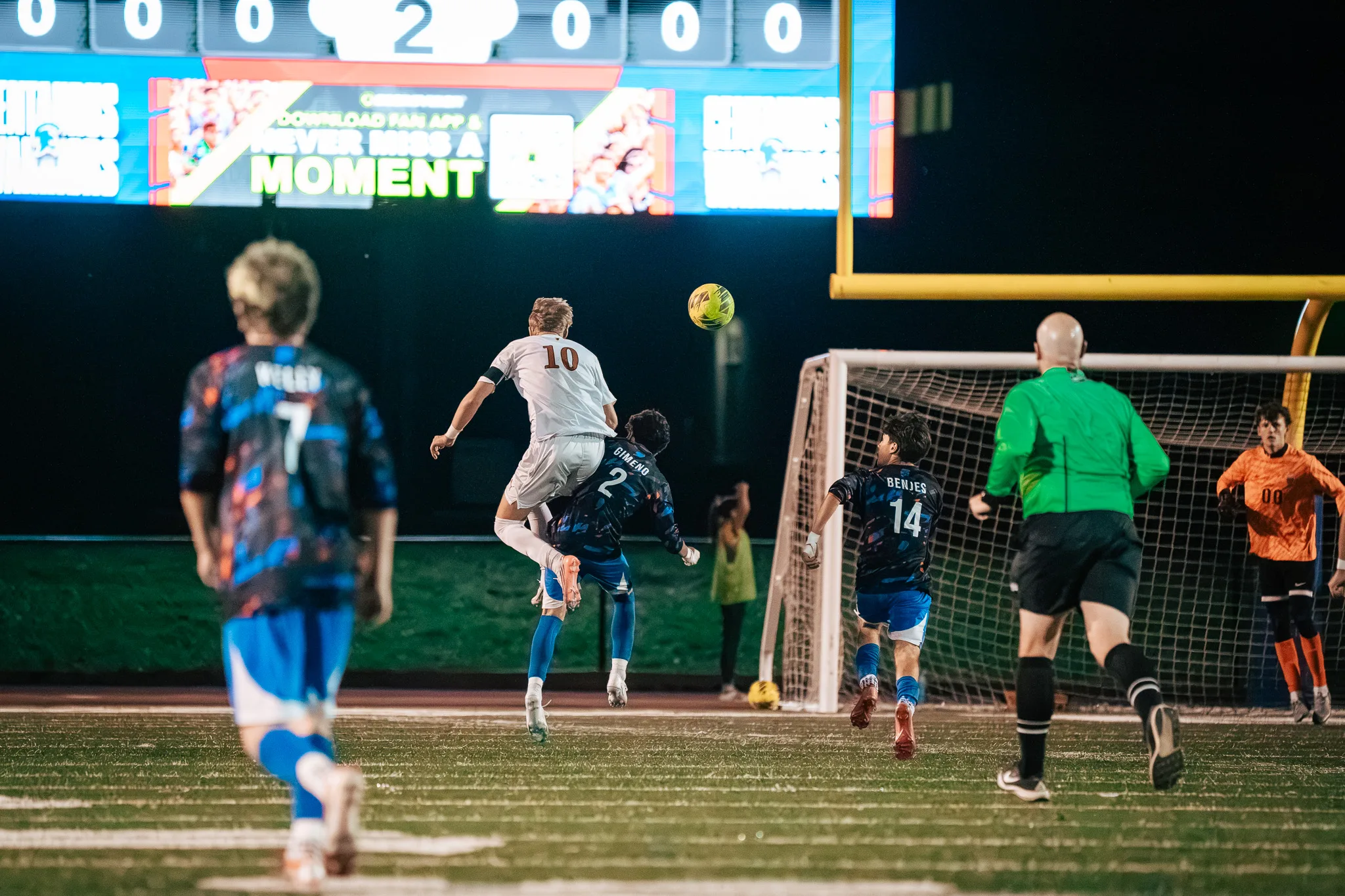 Mead number 10 and Centaurus number 2 leap together for a header as Centaurus number 14 runs toward the goal, where the goalkeeper in orange, number 00, and a referee in green wait, with Centaurus number 7 in the near foreground