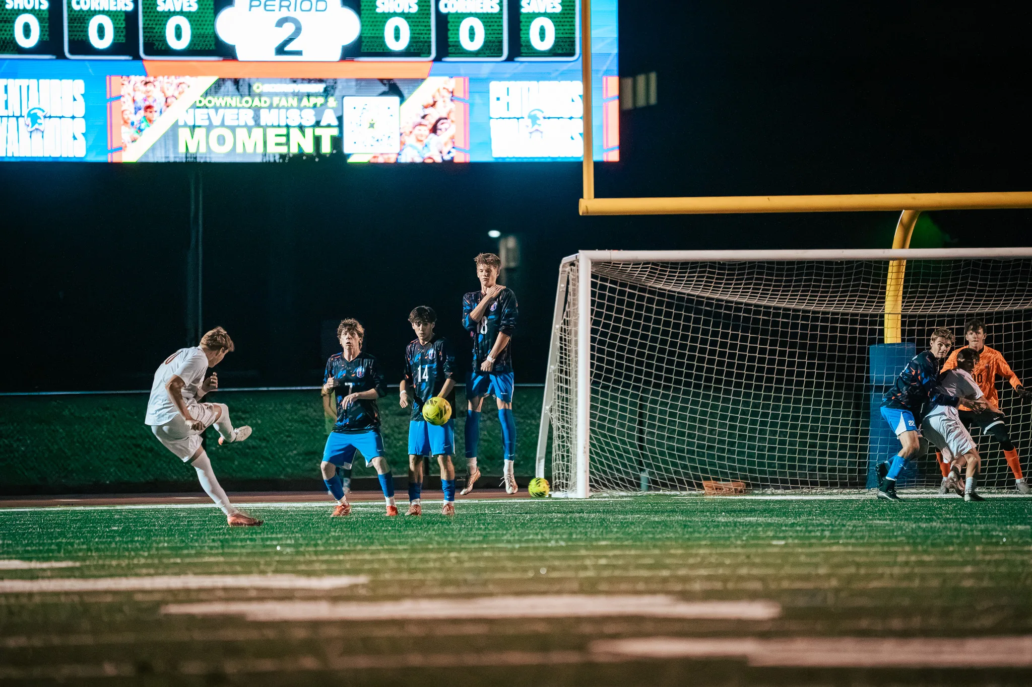 A Mead player follows through on a free kick as three Centaurus players stand in a wall with the ball in the air in front of them and players from both teams crowd the Centaurus goal at the right