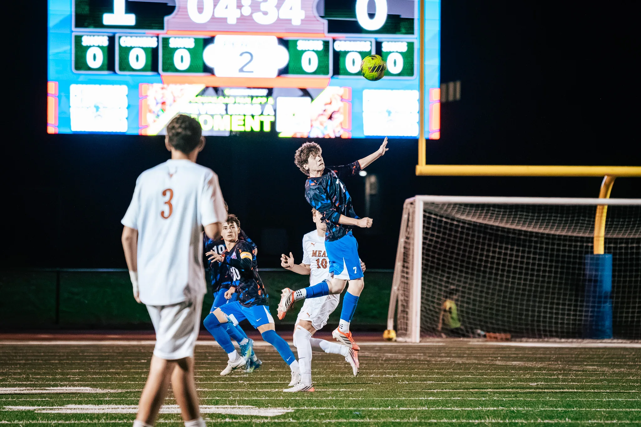 Centaurus number 7 leaps with an arm out to meet the ball as Mead number 10 rises beneath him and Mead number 3 watches from the foreground, the scoreboard behind them showing 1 to 0