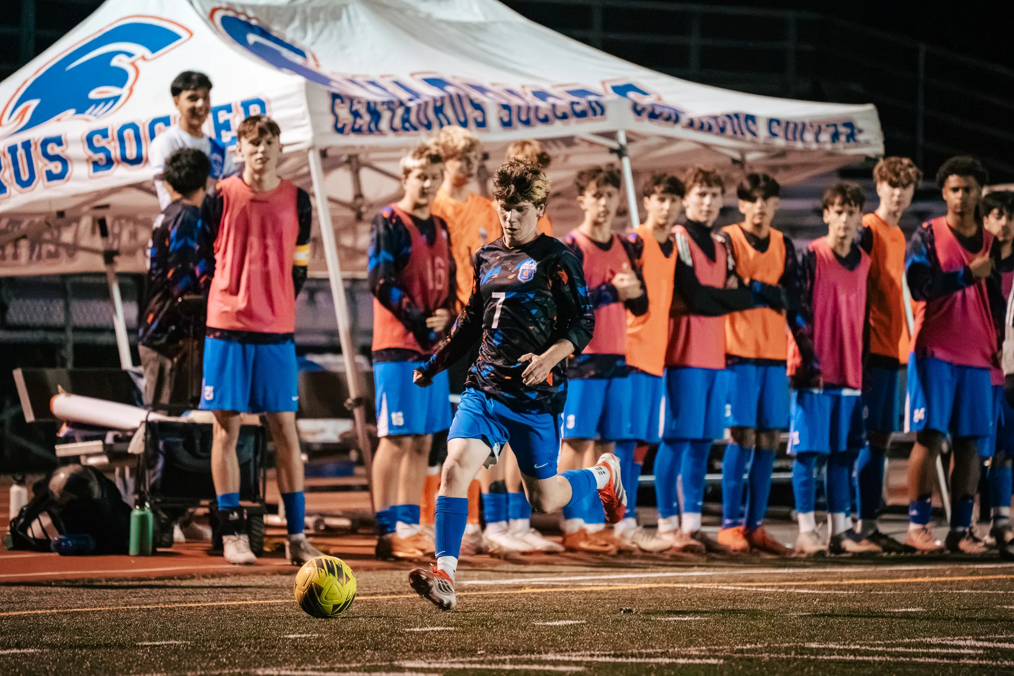Centaurus number 7 runs toward the ball in front of the team's white tent as a row of substitutes in orange and pink bibs watches from the sideline
