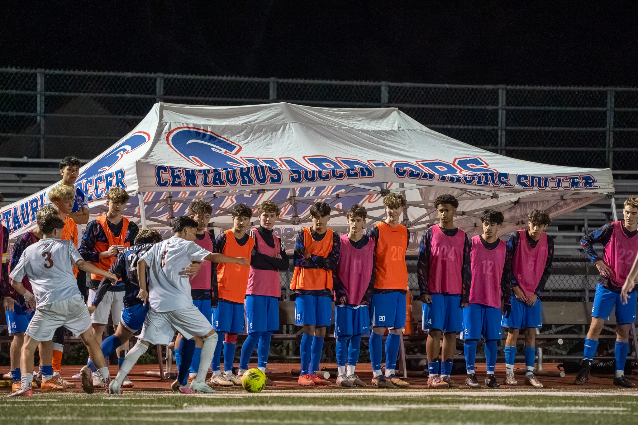 A row of Centaurus substitutes in orange and pink bibs stands under the team's tent, watching players from both teams battle for the ball at the left