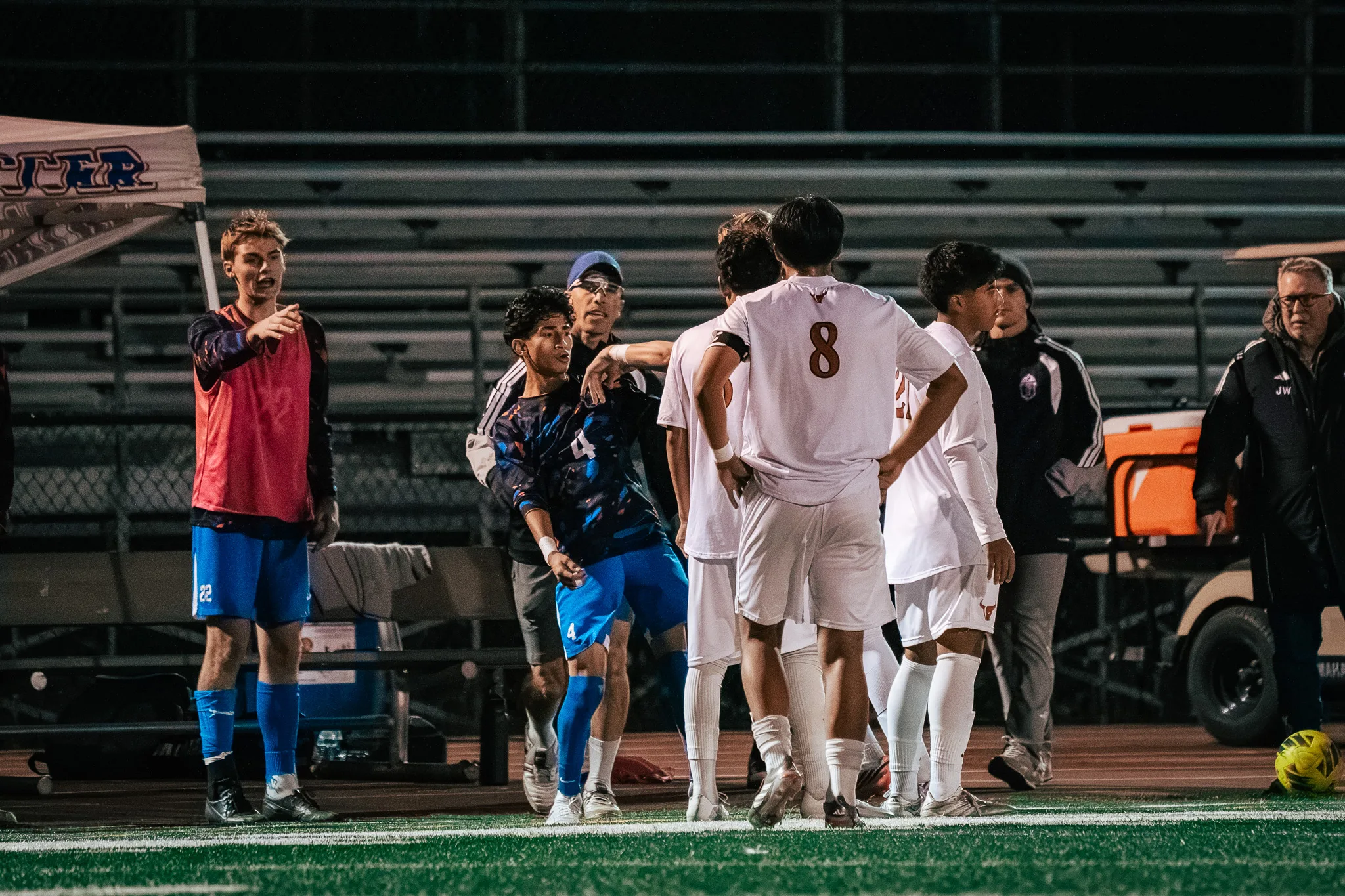 Near the Centaurus bench, a man in a blue cap and glasses puts an arm across Centaurus number 4 as Mead players, including number 8, stand close, while a Centaurus substitute in a red bib, number 22, points toward the field