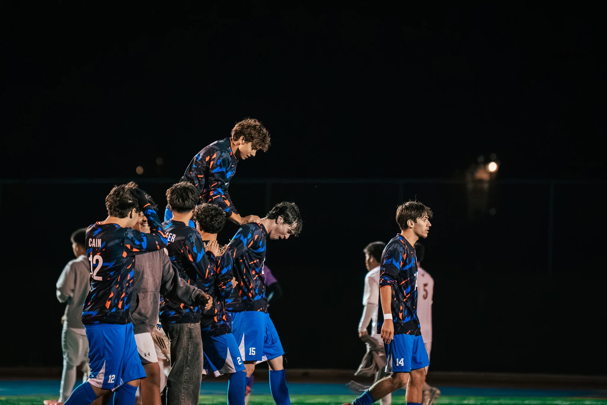 Centaurus players pile together at the edge of the field, one climbing above the group with a hand on a teammate's head, while number 14 walks past at the right and Mead players stand in the background