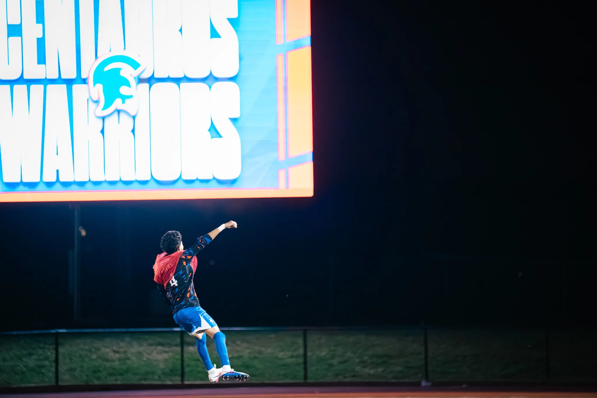 Centaurus number 4, a red bib bunched at his neck, jumps with a raised fist toward the stadium scoreboard, which reads Centaurus Warriors