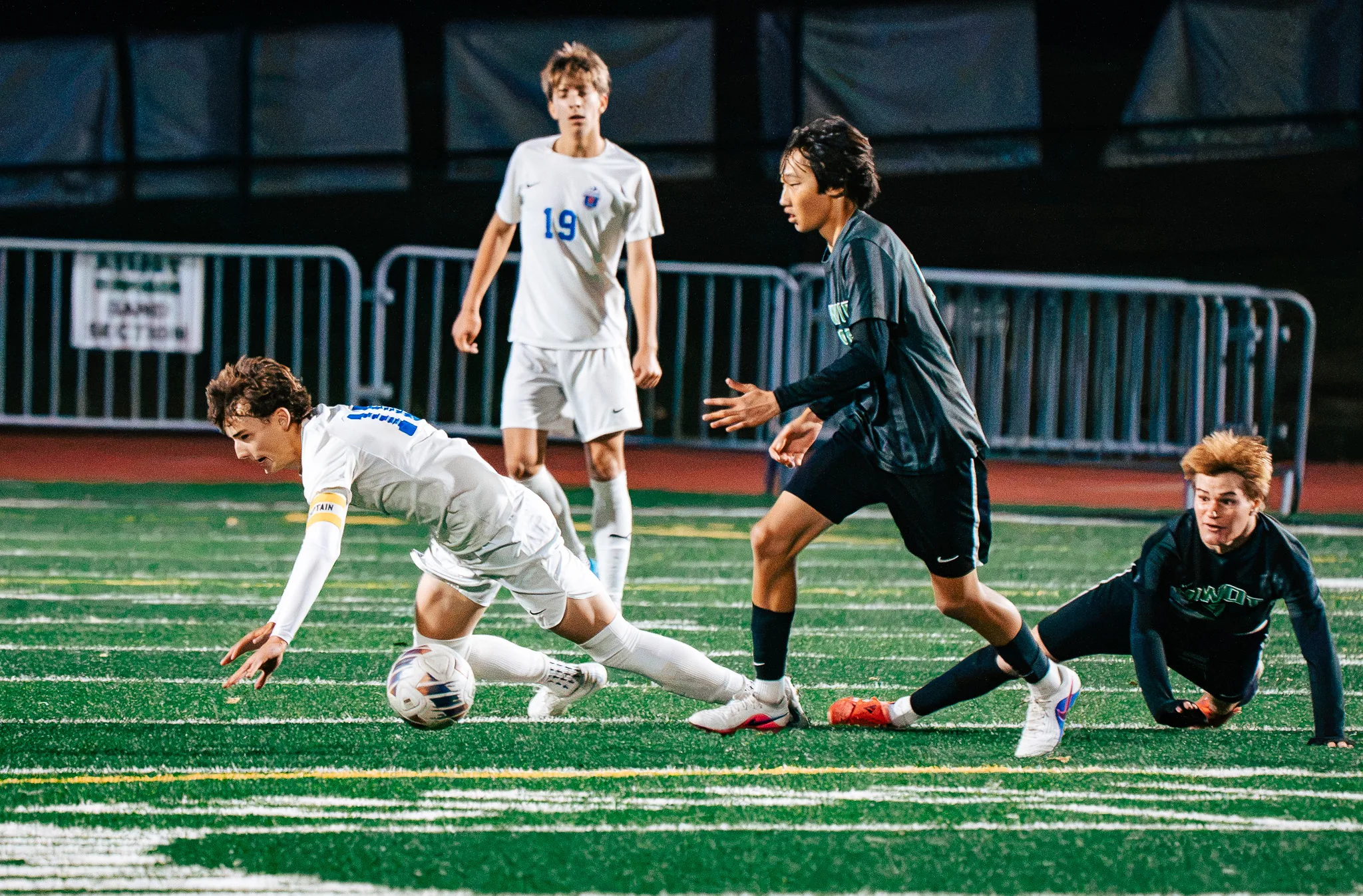 A Centaurus player in a captain's armband stumbles forward over the ball between two Niwot players, one sprawled on the turf, as Centaurus number 19 watches behind