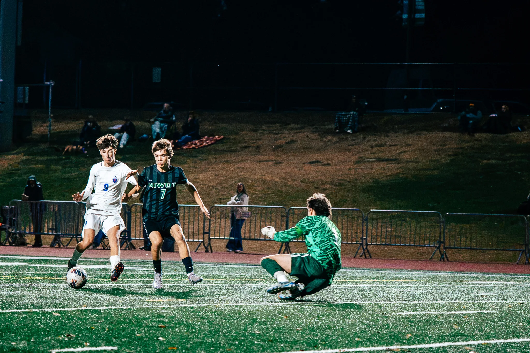 Centaurus number 9 brings the ball in as Niwot number 7 runs beside him and the Niwot goalkeeper in green goes down to block, spectators on the hill behind