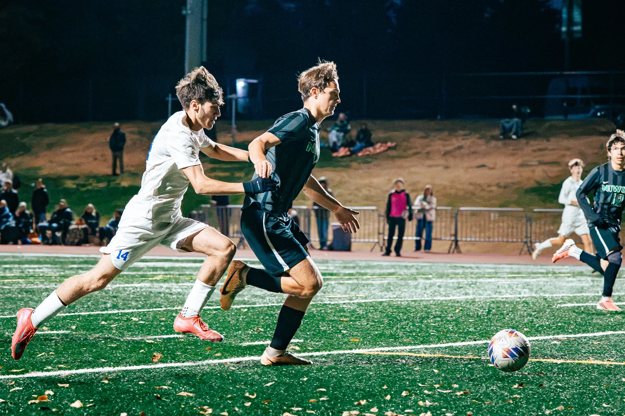 Centaurus number 14 chases a Niwot player toward the ball with a hand on his back, spectators lining the hill behind