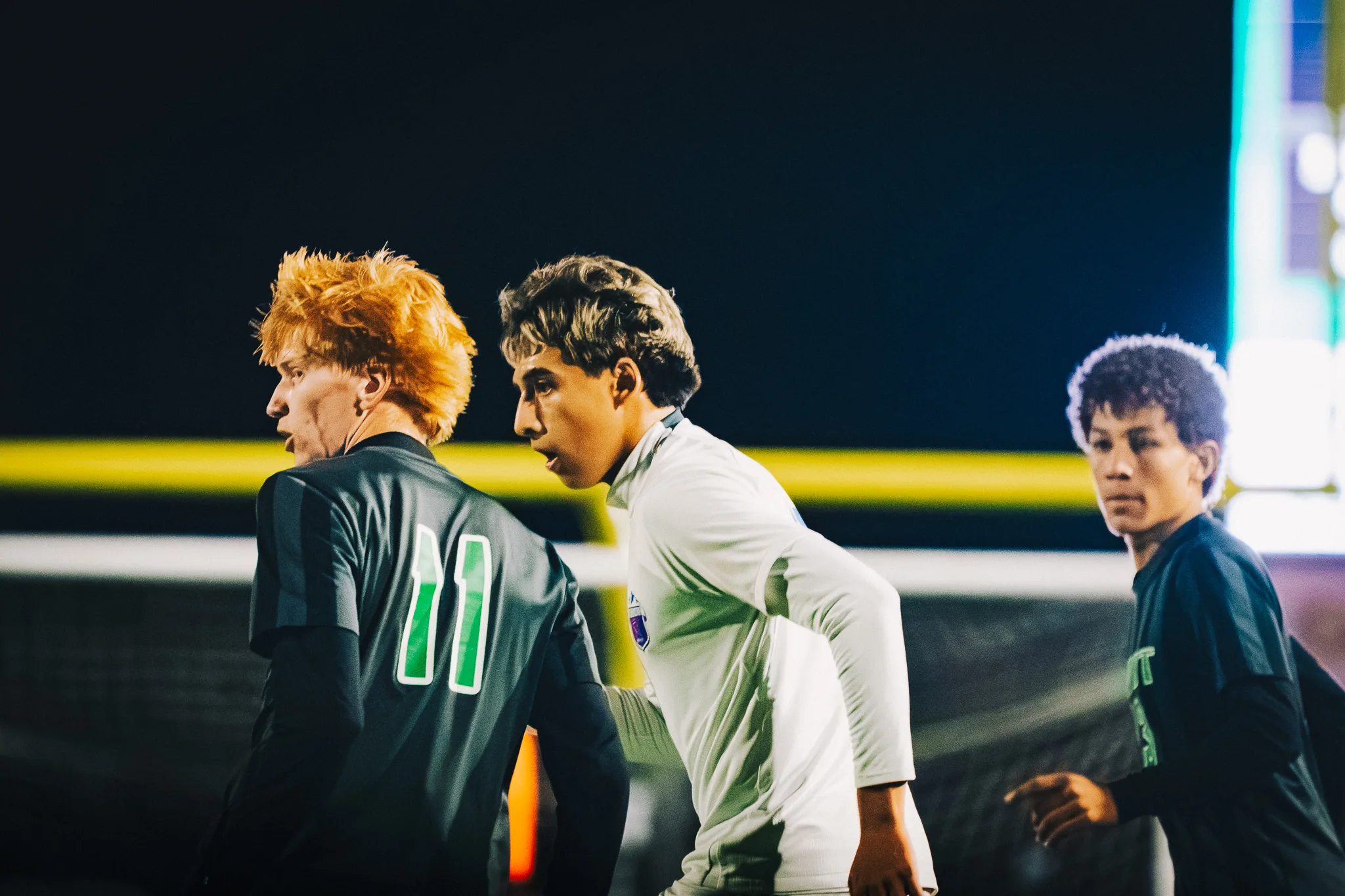 Niwot number 11, with red hair, and a Centaurus player stand side by side watching the play, a Niwot teammate pointing at the right