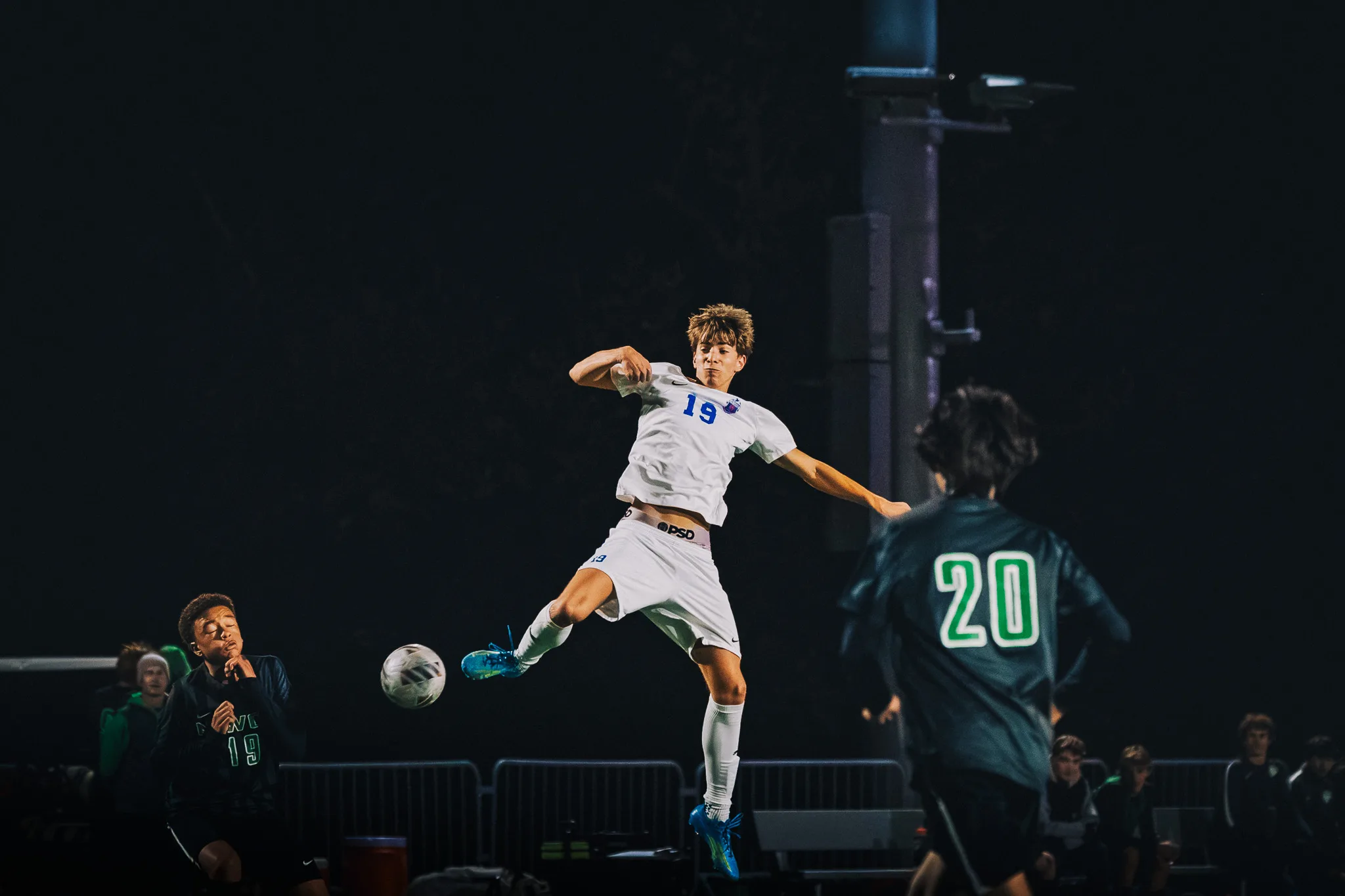 Centaurus number 19 hangs in the air with his leg swung up toward the ball as Niwot number 20 runs in and a Niwot player on the sideline watches with his hands at his chin