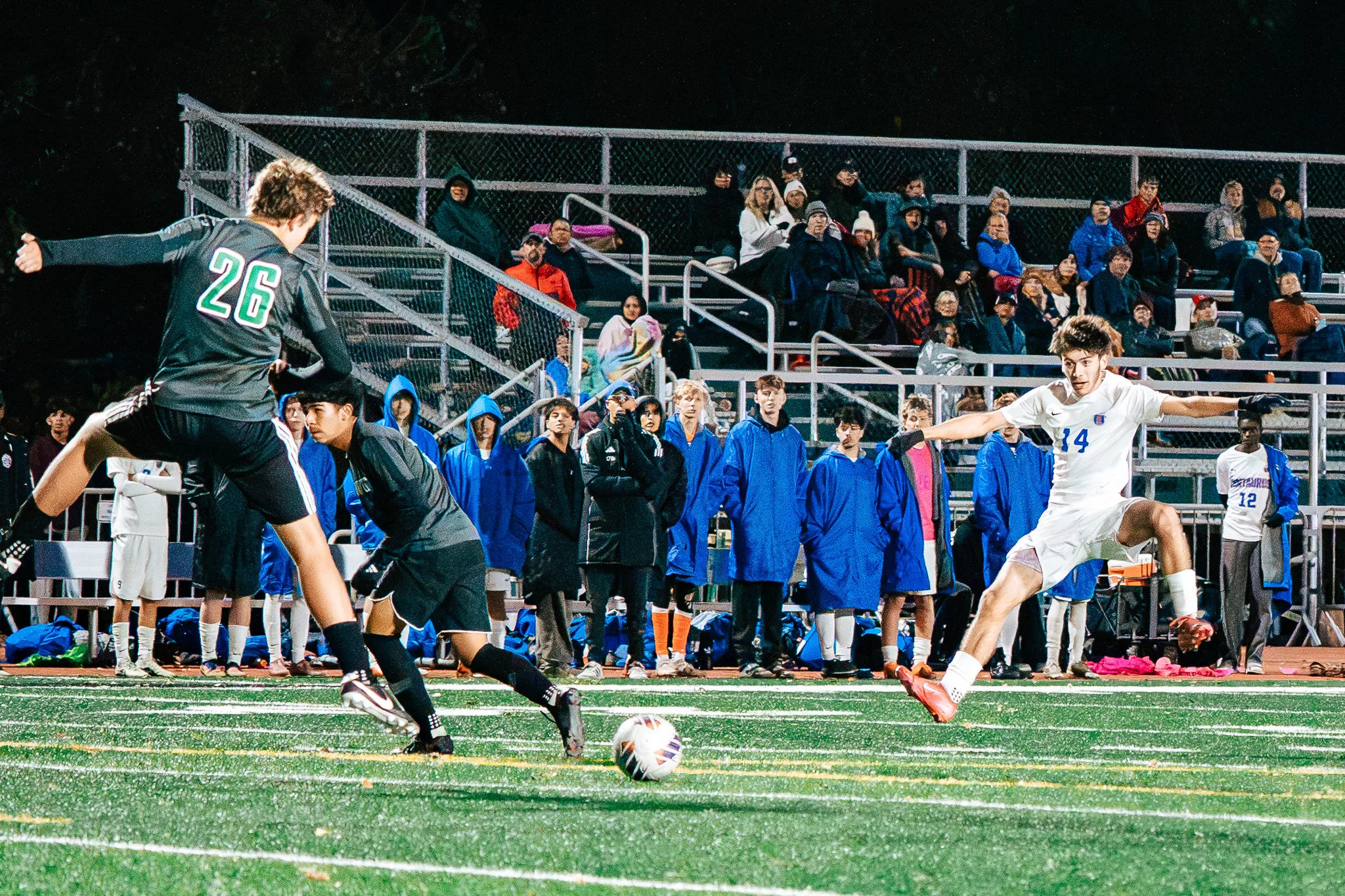 Niwot number 26 winds up to clear the ball as Centaurus number 14 lunges in with his arms spread, the Centaurus bench in blue parkas and the packed bleachers behind