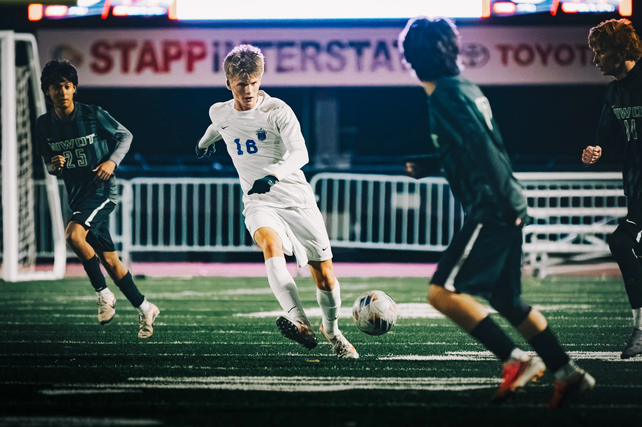 Centaurus number 18 dribbles into a gap between Niwot number 25 and two more Niwot players, a lit sponsor banner above the fence