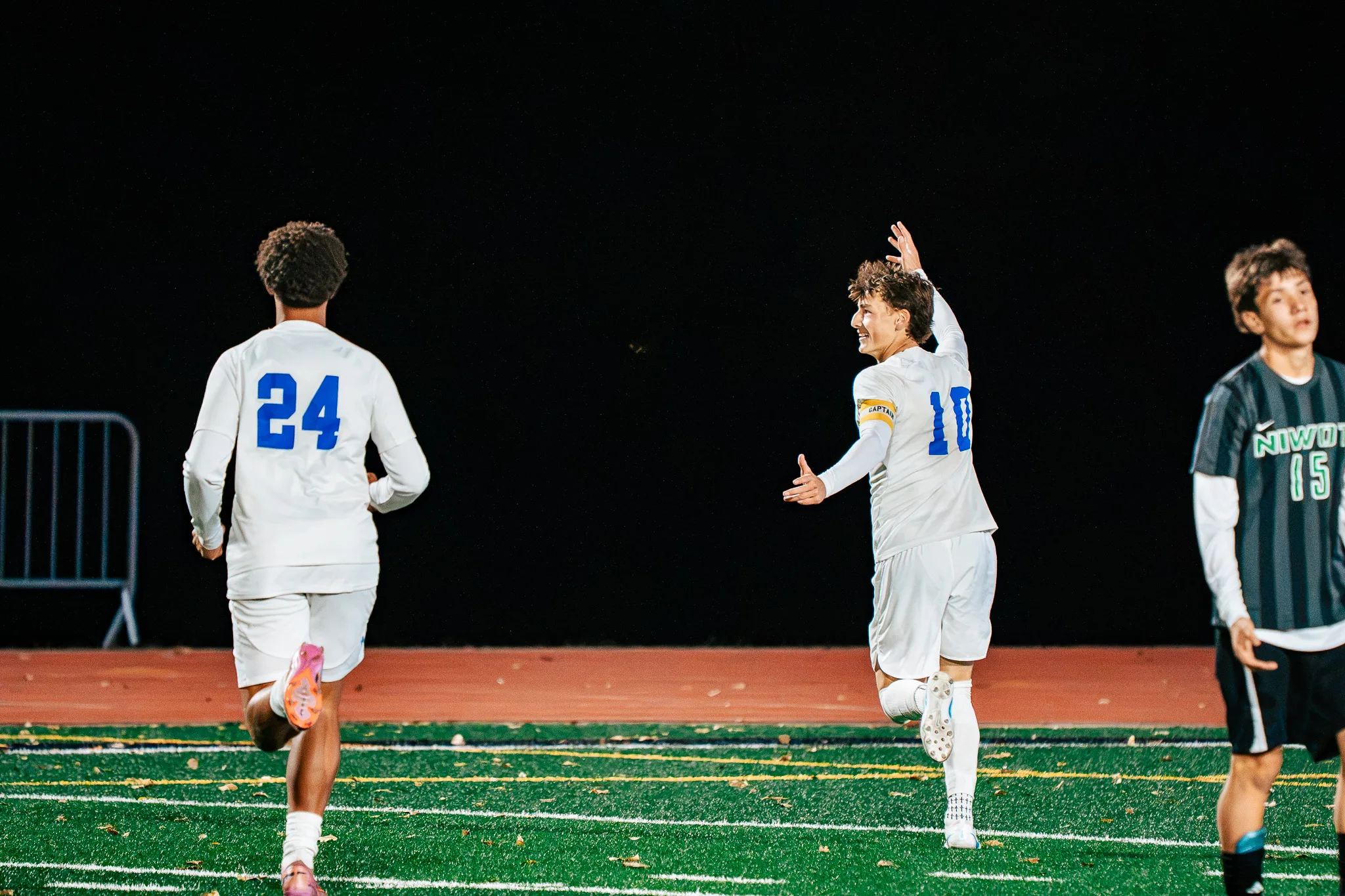 Smiling, Centaurus number 10 in a captain's armband raises a hand and looks back at his teammates as number 24 runs ahead and Niwot number 15 walks away