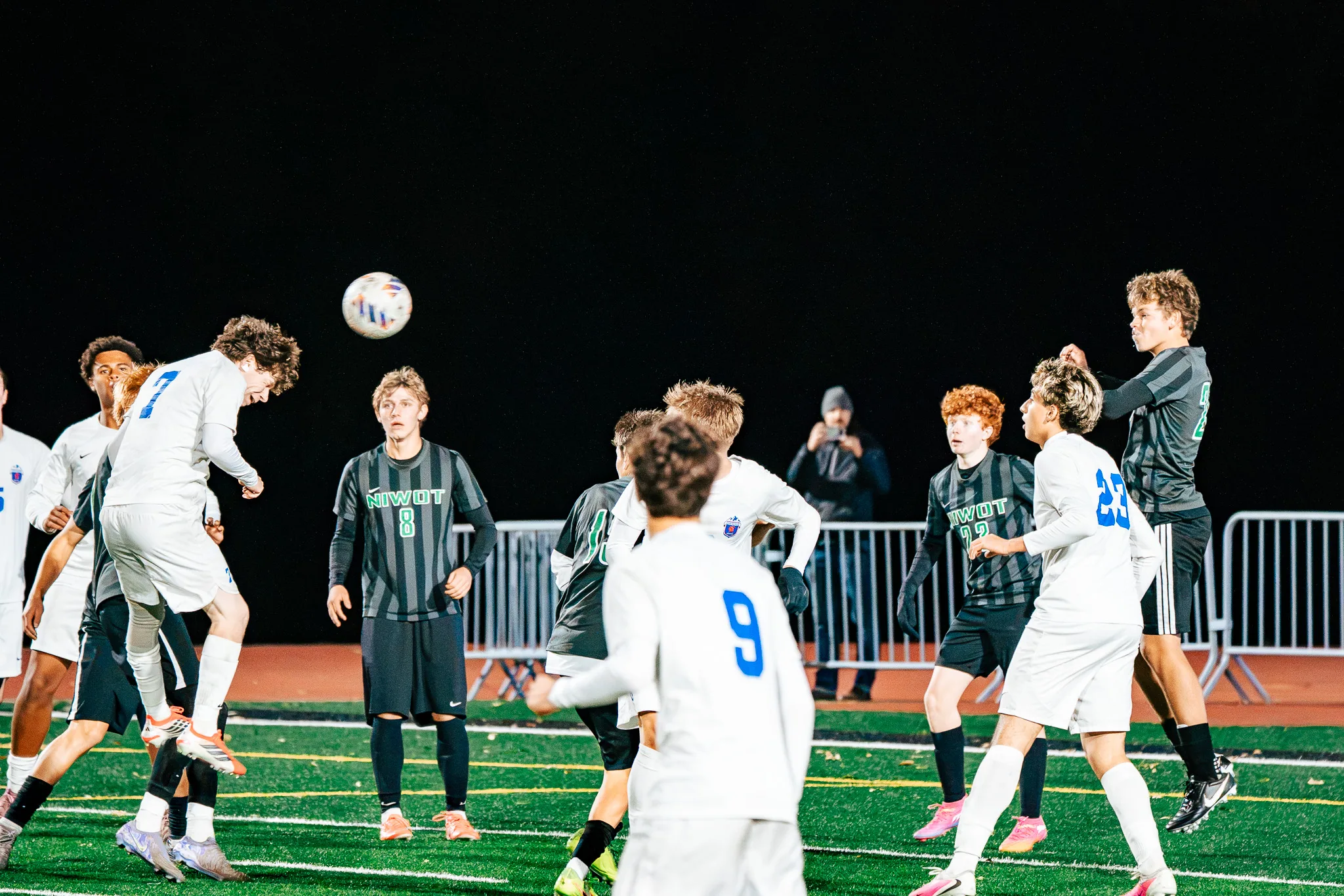A Centaurus player rises to head the ball in a crowd of players as Niwot number 8 watches and Centaurus numbers 9 and 23 look on from the foreground, a fan holding up a phone behind the fence