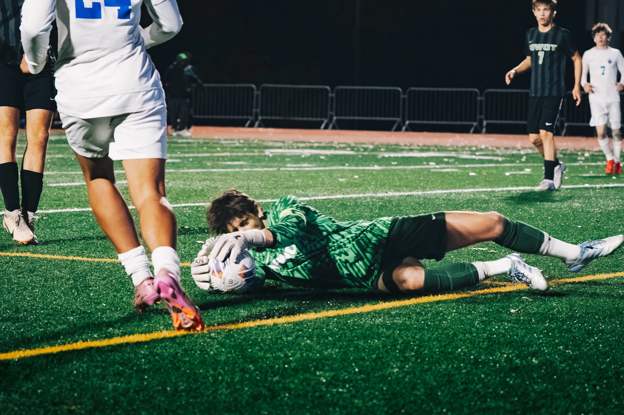 The Niwot goalkeeper in green lies on his side hugging the ball to his chest as Centaurus number 24 steps past, Niwot number 7 and Centaurus number 7 watching behind