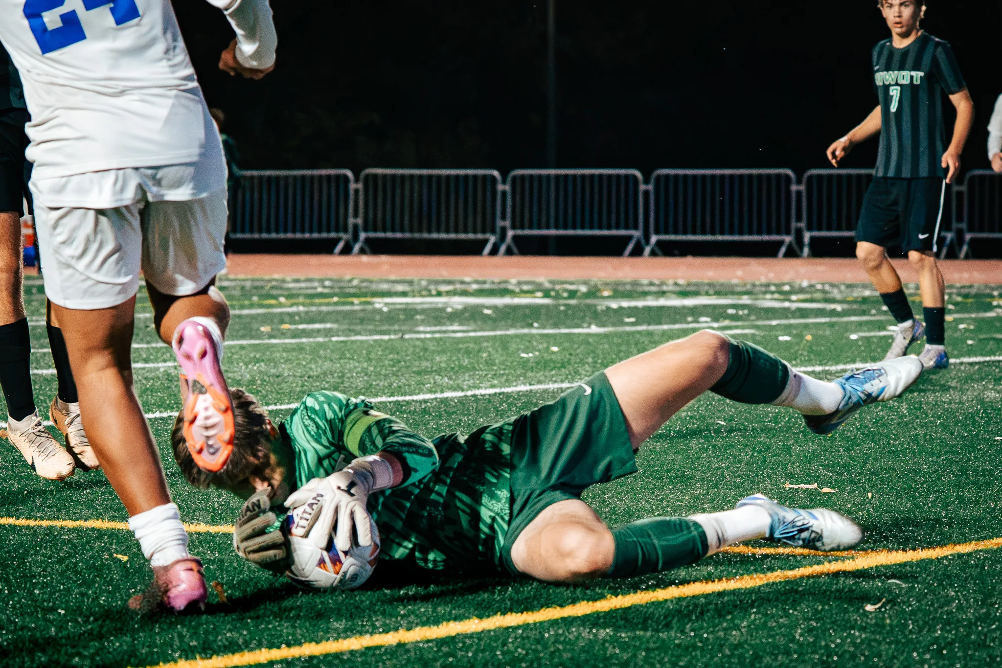The Niwot goalkeeper curls around the ball on the turf as Centaurus number 24 steps over him, Niwot number 7 jogging in behind