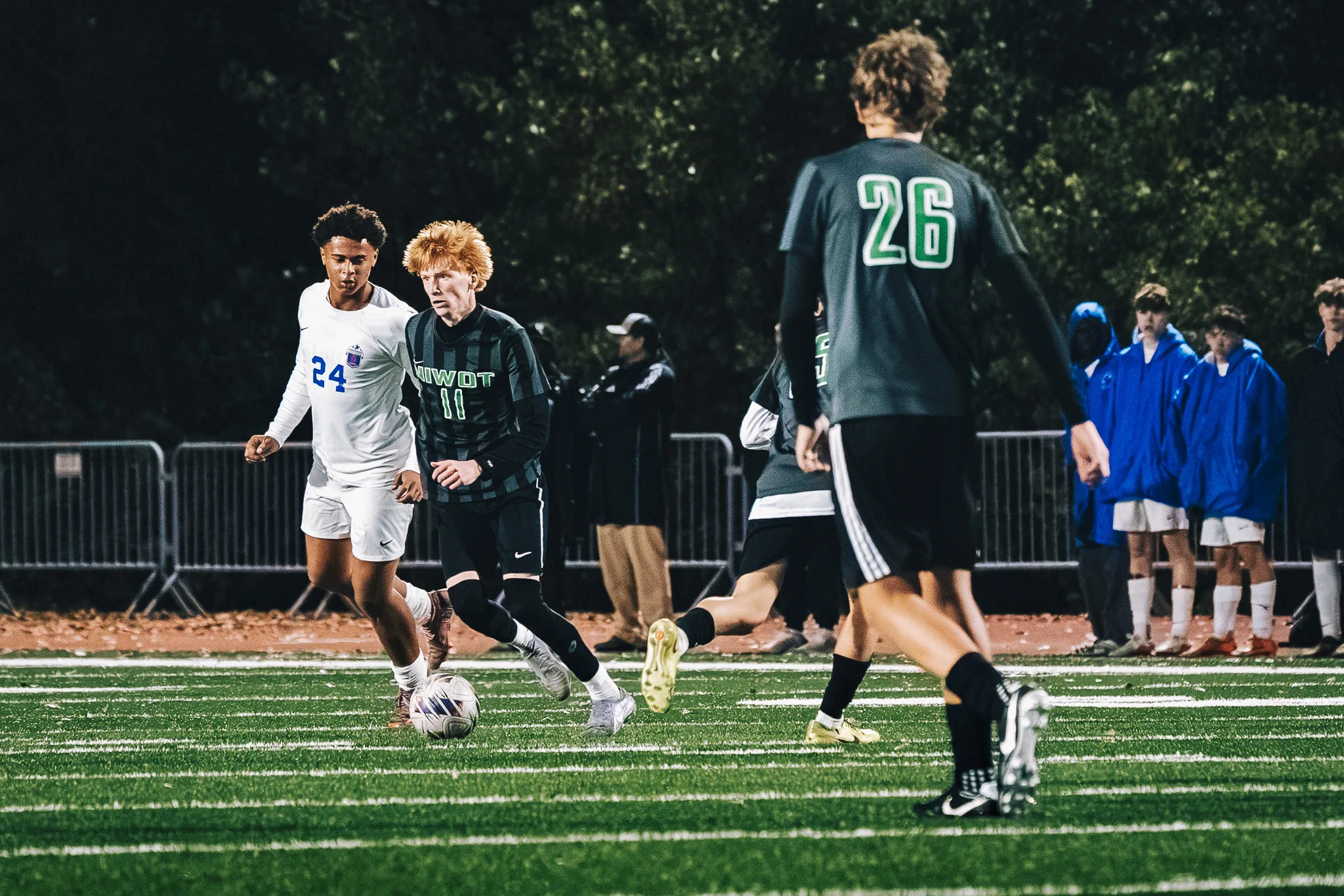 Niwot number 11 dribbles away from Centaurus number 24 as Niwot number 26 looks on, Centaurus substitutes in blue parkas watching from the sideline