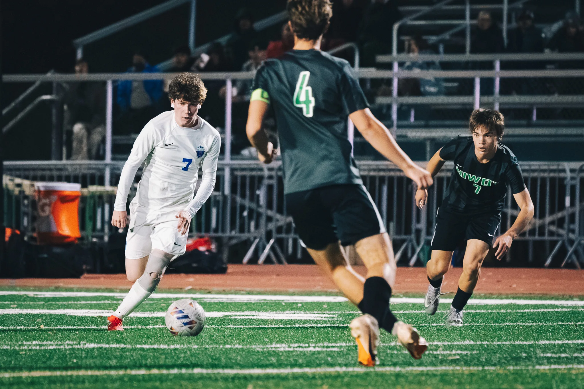 Centaurus number 7 dribbles toward Niwot number 4, who wears a green armband, as Niwot number 7 closes in, spectators in the bleachers behind
