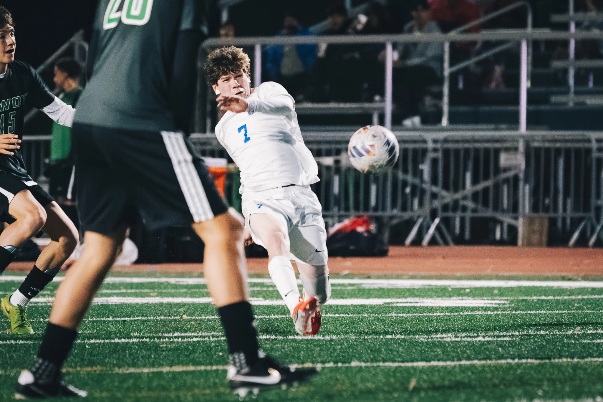 Centaurus number 7 follows through on a kick as the ball flies away, a Niwot player blurred in the foreground and the bleachers behind