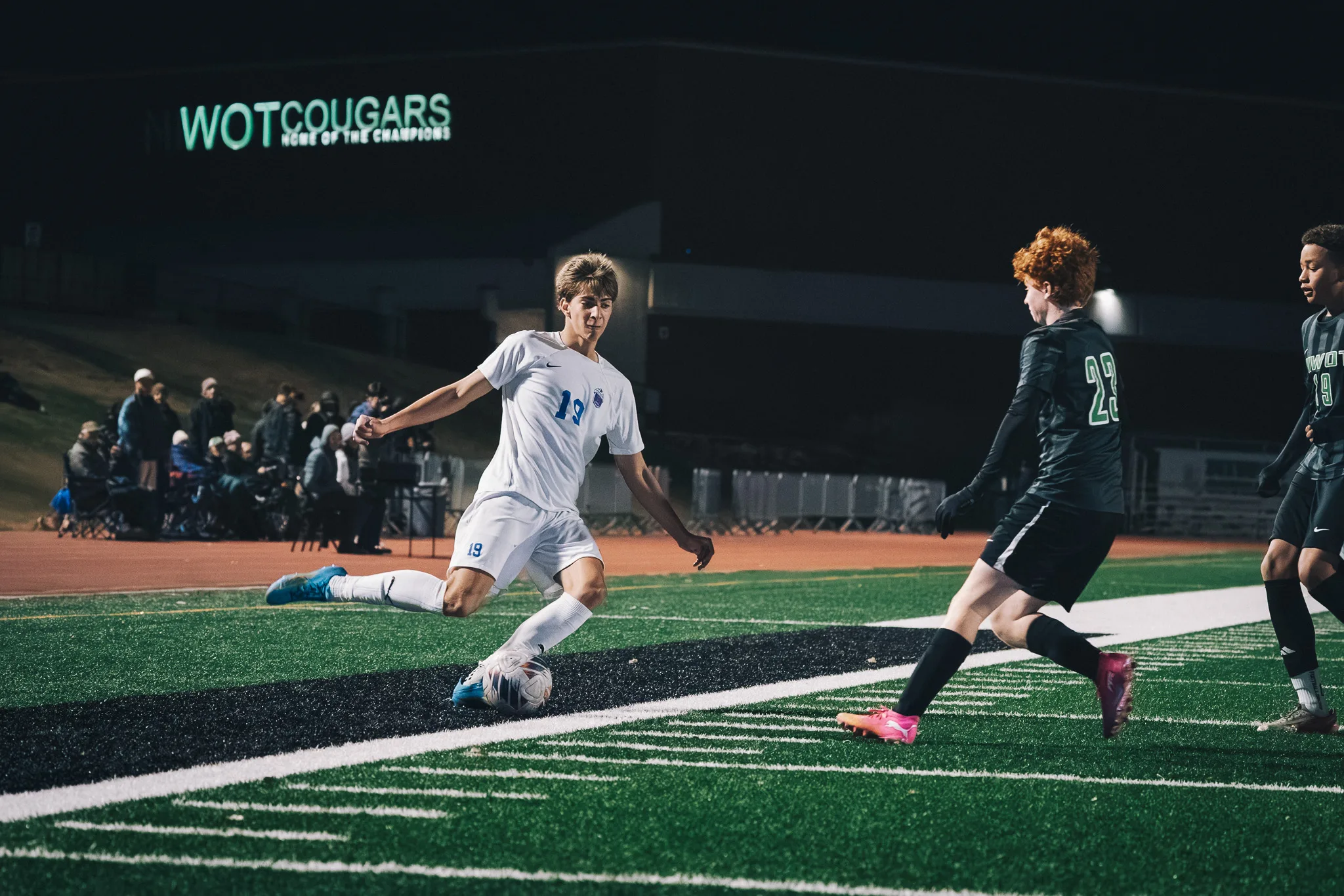 Centaurus number 19 swings his leg through the ball near the sideline as a Niwot player with red hair closes in, a lit Niwot Cougars sign on the building behind