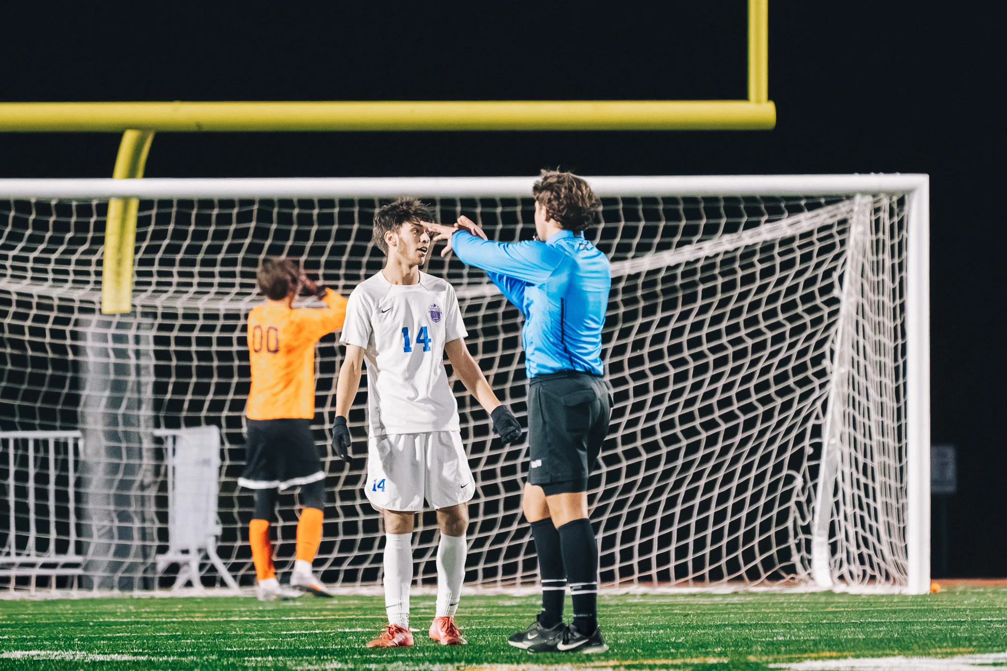 In front of the goal, a referee in blue gestures as he talks with Centaurus number 14, the Centaurus goalkeeper in orange, number 00, behind them