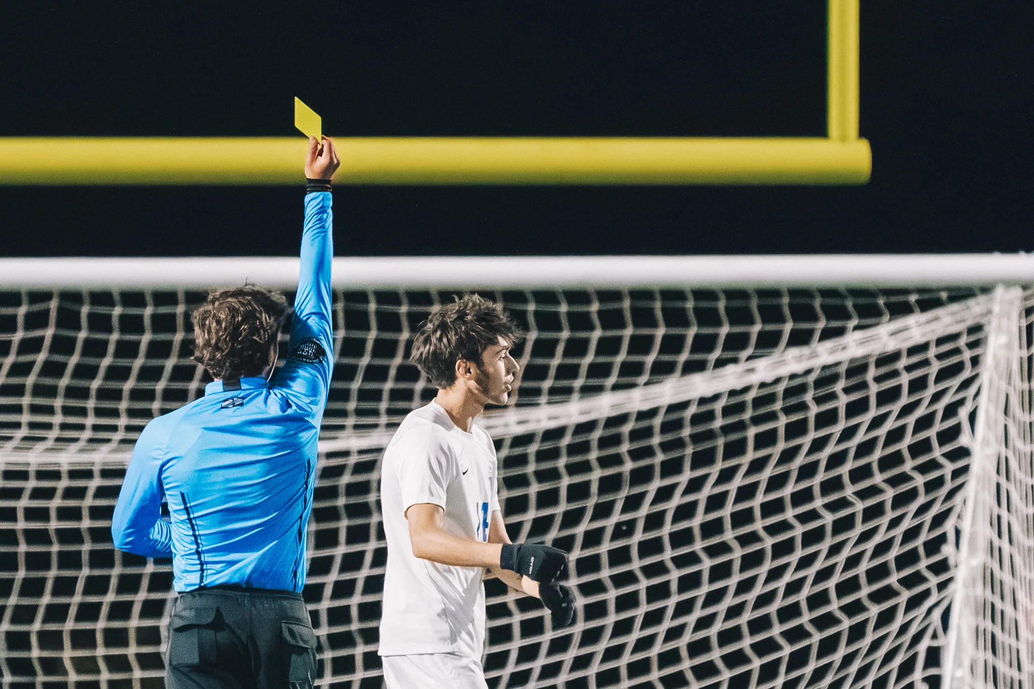 In front of the goal, a referee in blue holds up a yellow card as a Centaurus player in gloves looks away