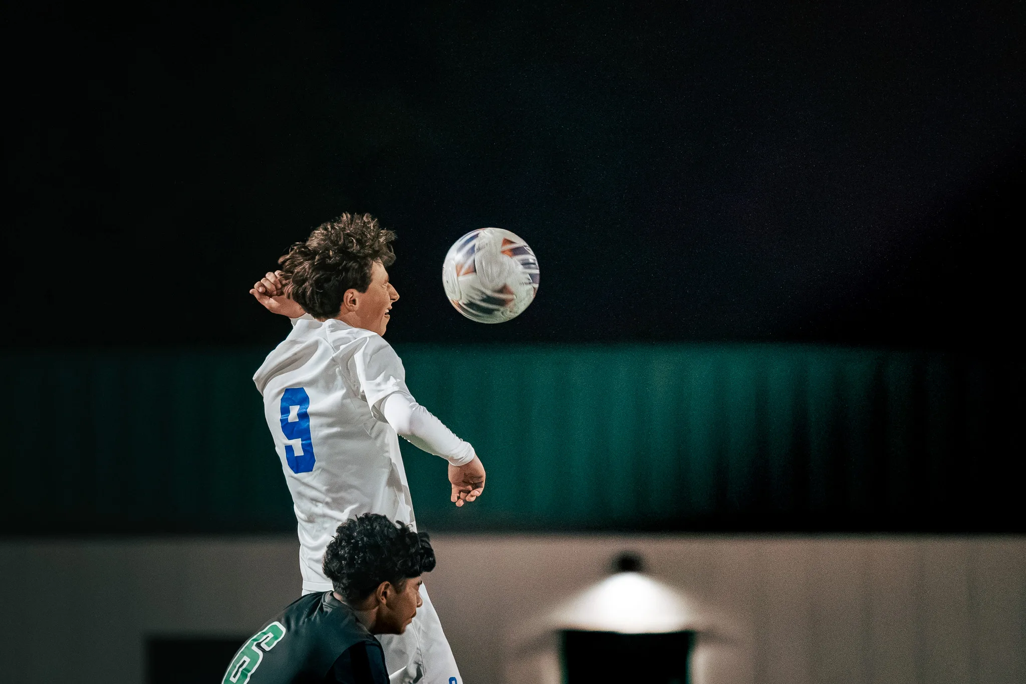 Centaurus number 9 rises to head the ball at night, a Niwot player looking up from below