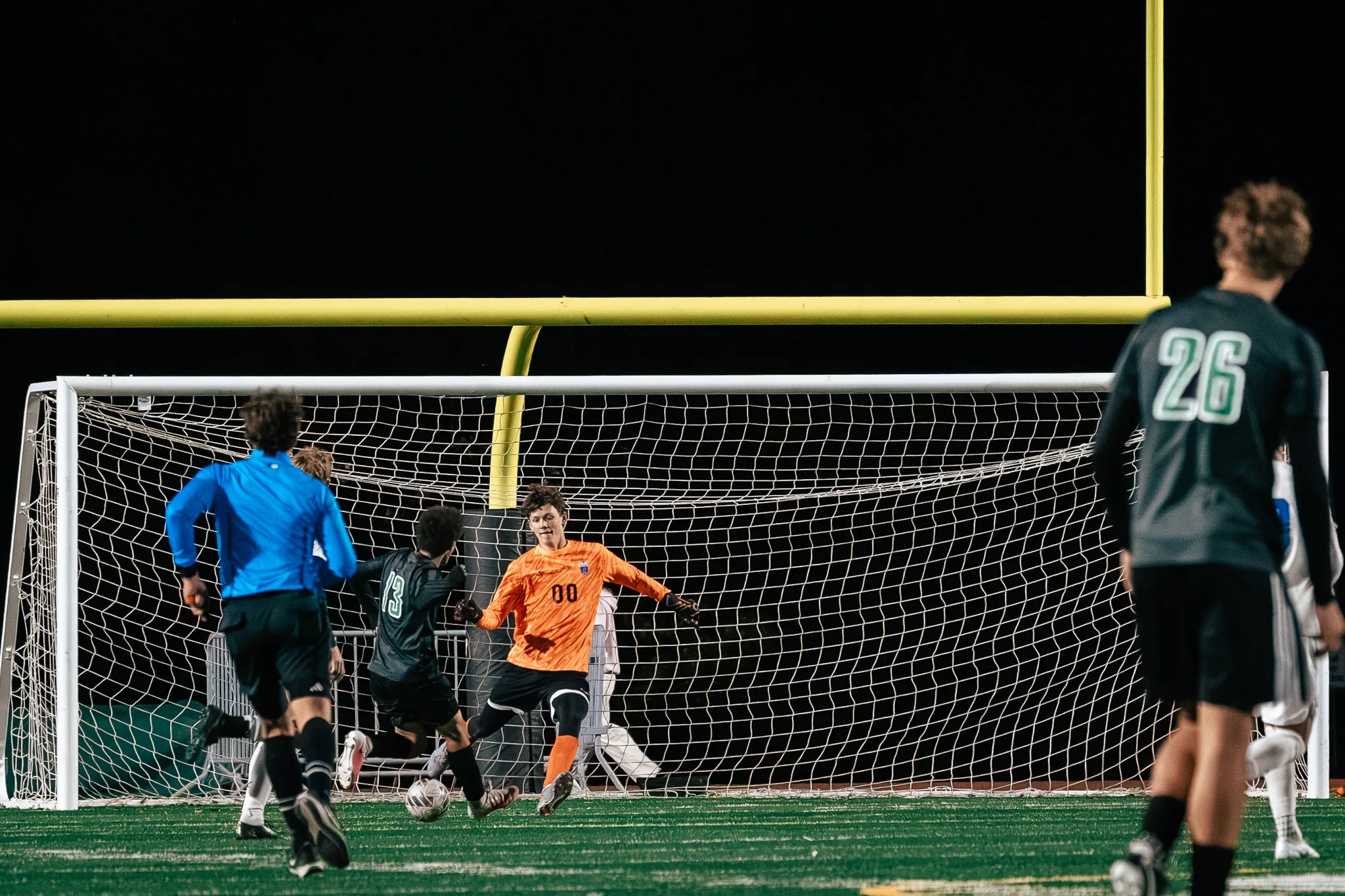 Niwot number 13 strikes the ball at the goal as the Centaurus goalkeeper in orange, number 00, spreads to block, Niwot number 26 and the referee in blue watching