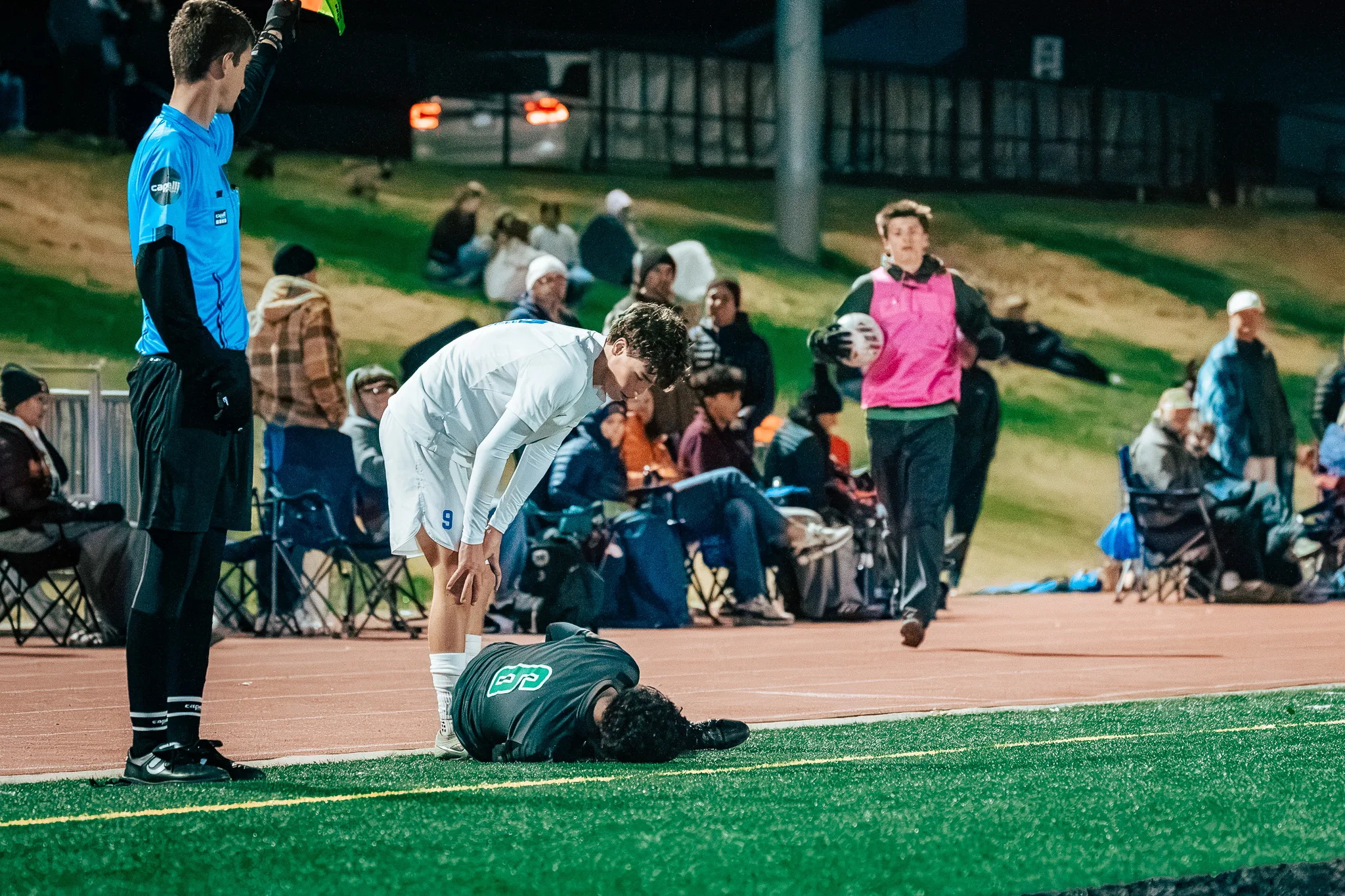 Centaurus number 9 bends over a Niwot player lying face down on the turf as an assistant referee waits beside them, a substitute in a pink bib jogging over with a ball