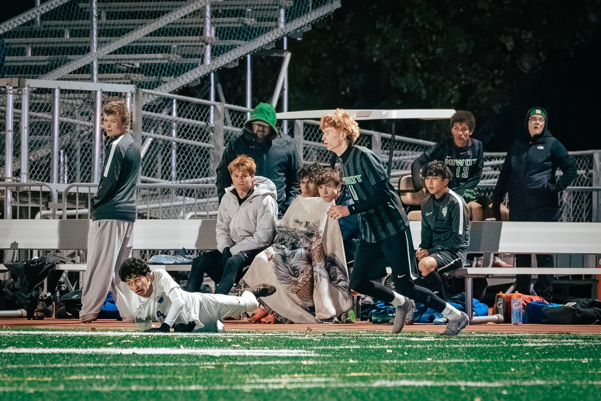 A Centaurus player slides across the turf in front of the Niwot bench as Niwot number 11, with red hair, runs past and players on the bench, one under a blanket, look on
