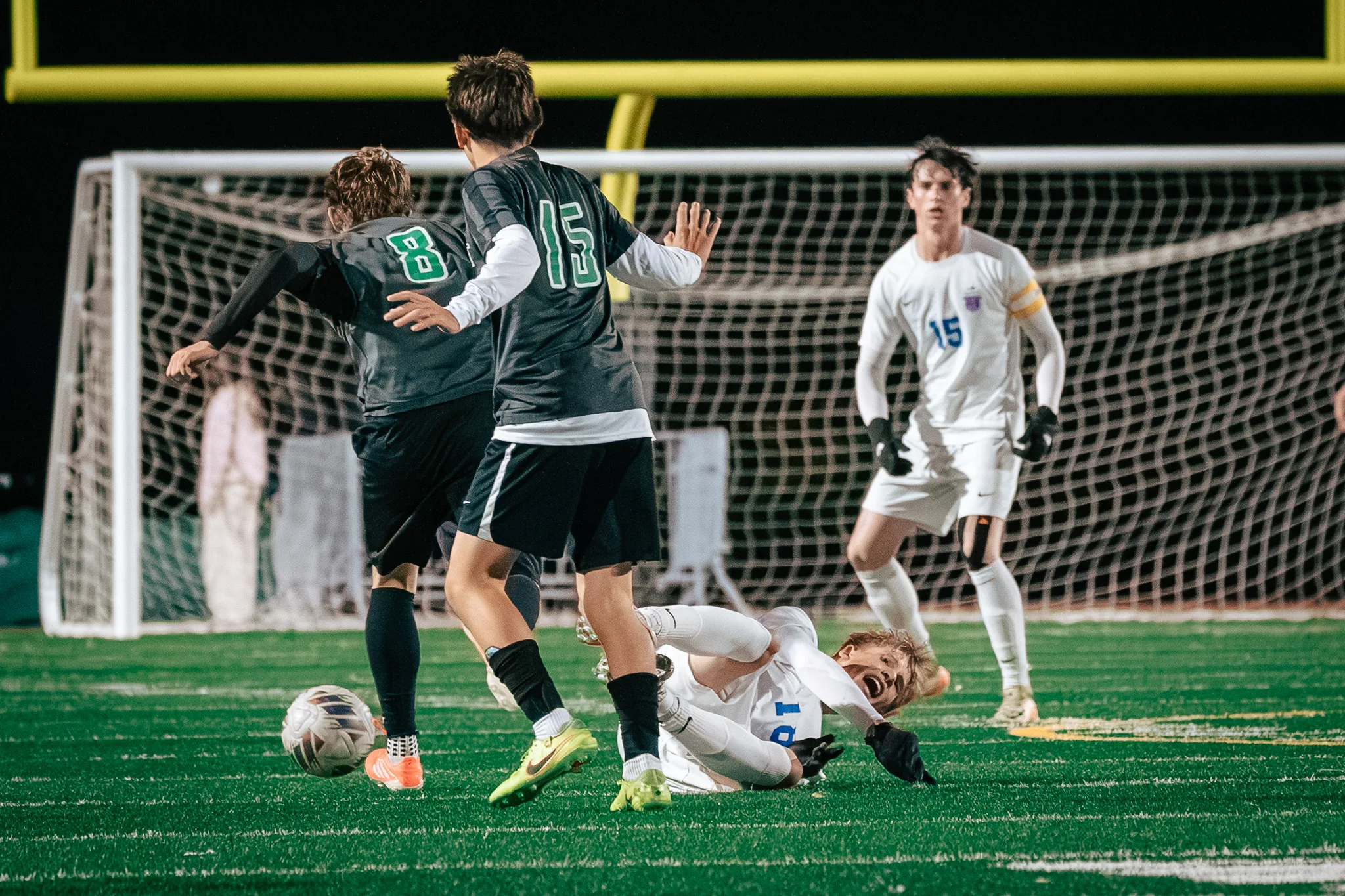 A Centaurus player lies on the turf with his mouth open as Niwot numbers 8 and 15 run on with the ball and Centaurus number 15 looks on in front of the goal