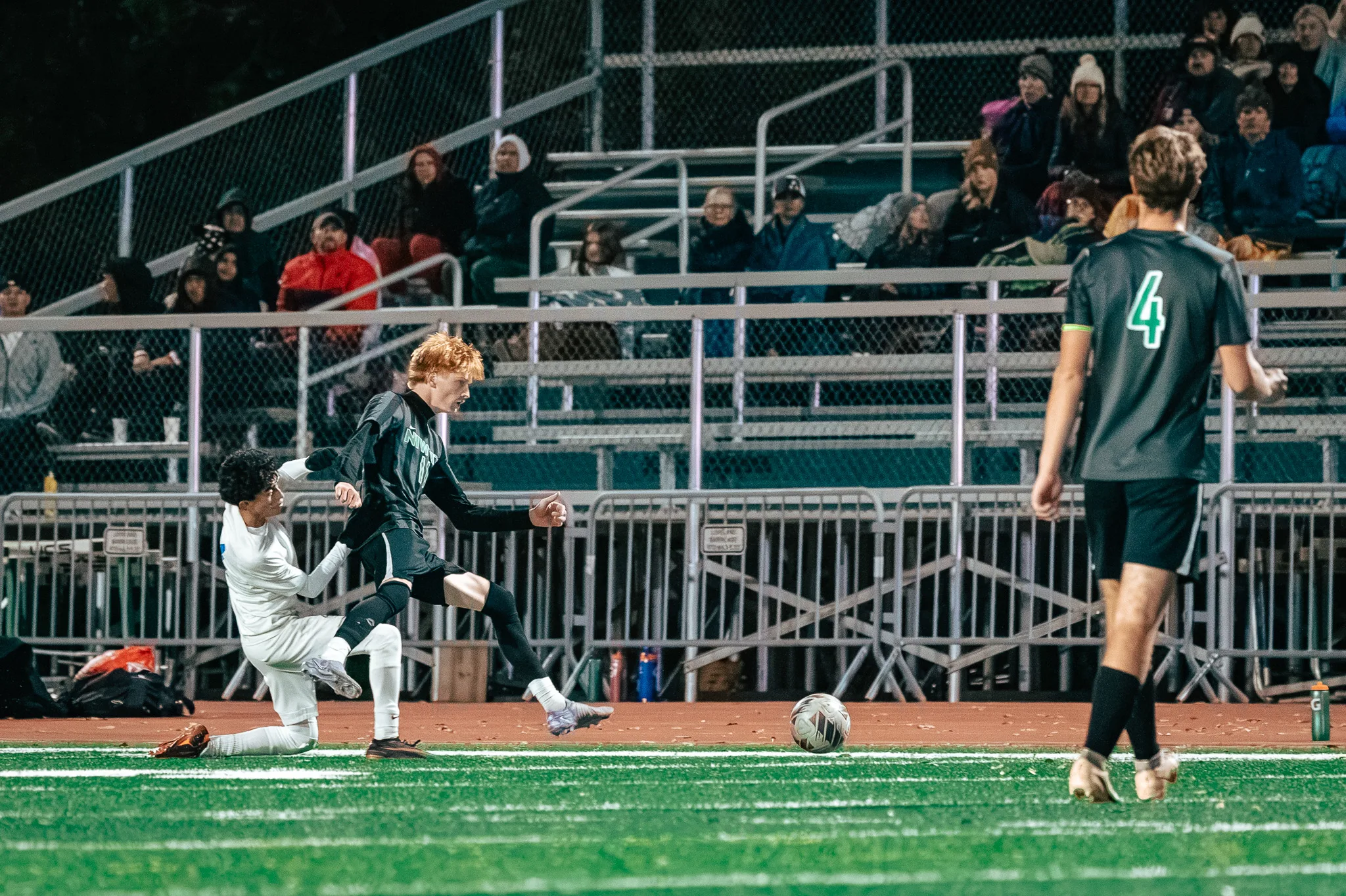 In front of the packed bleachers, a Niwot player with red hair leaps past a Centaurus player down on one knee as the ball rolls away and Niwot number 4 looks on